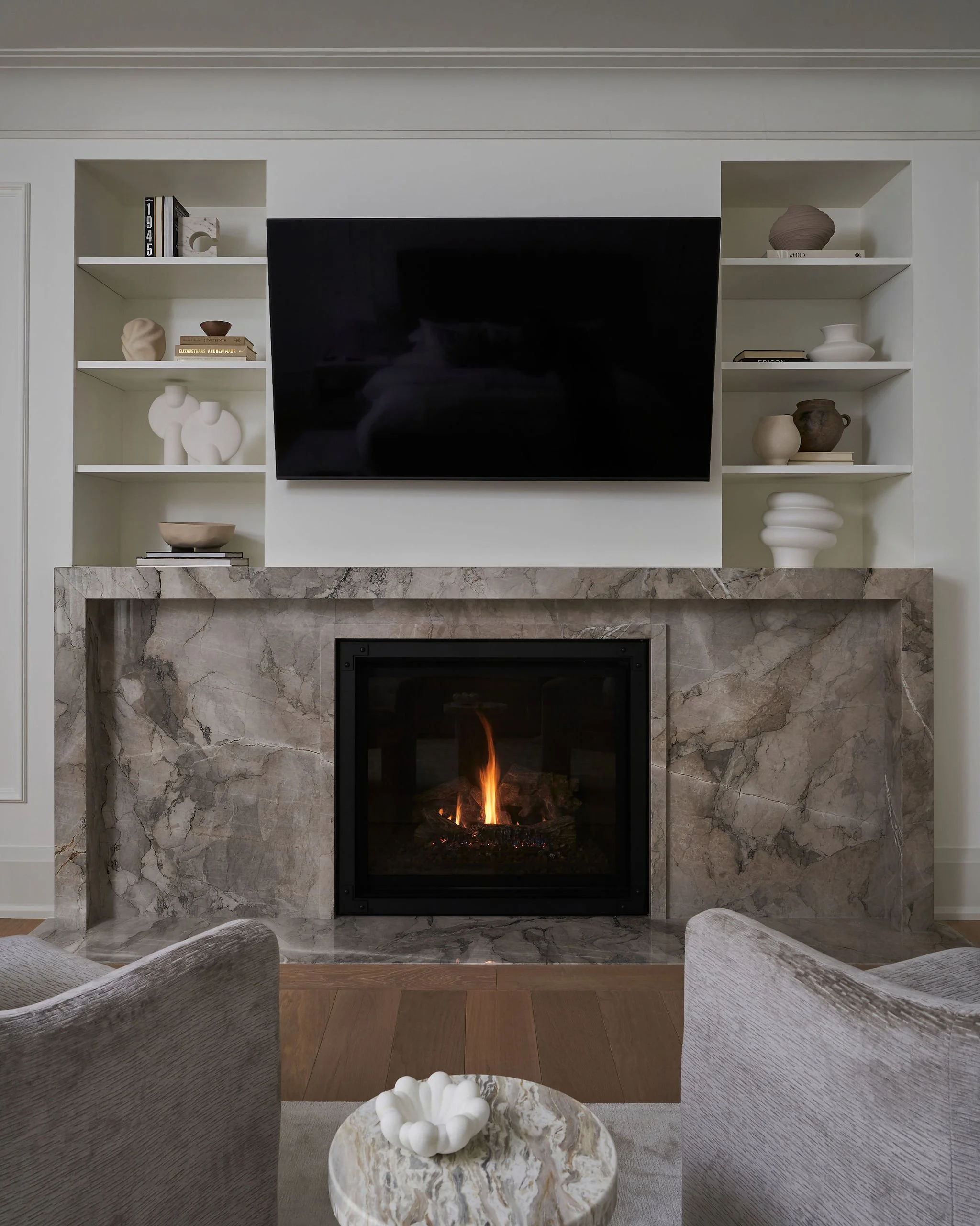 Living room with a built-in fireplace, white shelves with decorative objects, and a wall-mounted flat-screen TV.