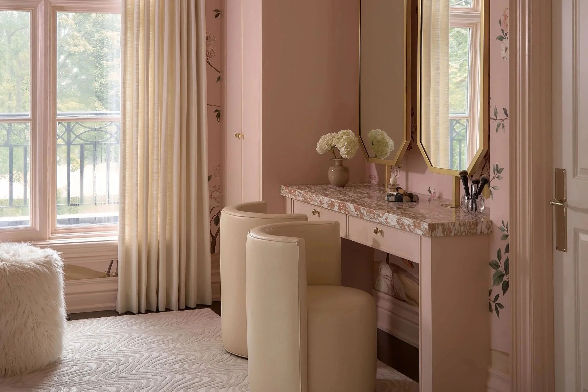 A cozy hotel room with large windows, cream curtains, a vanity table with two mirrors, white chairs, floral wallpaper, and a fluffy white chair in the corner.