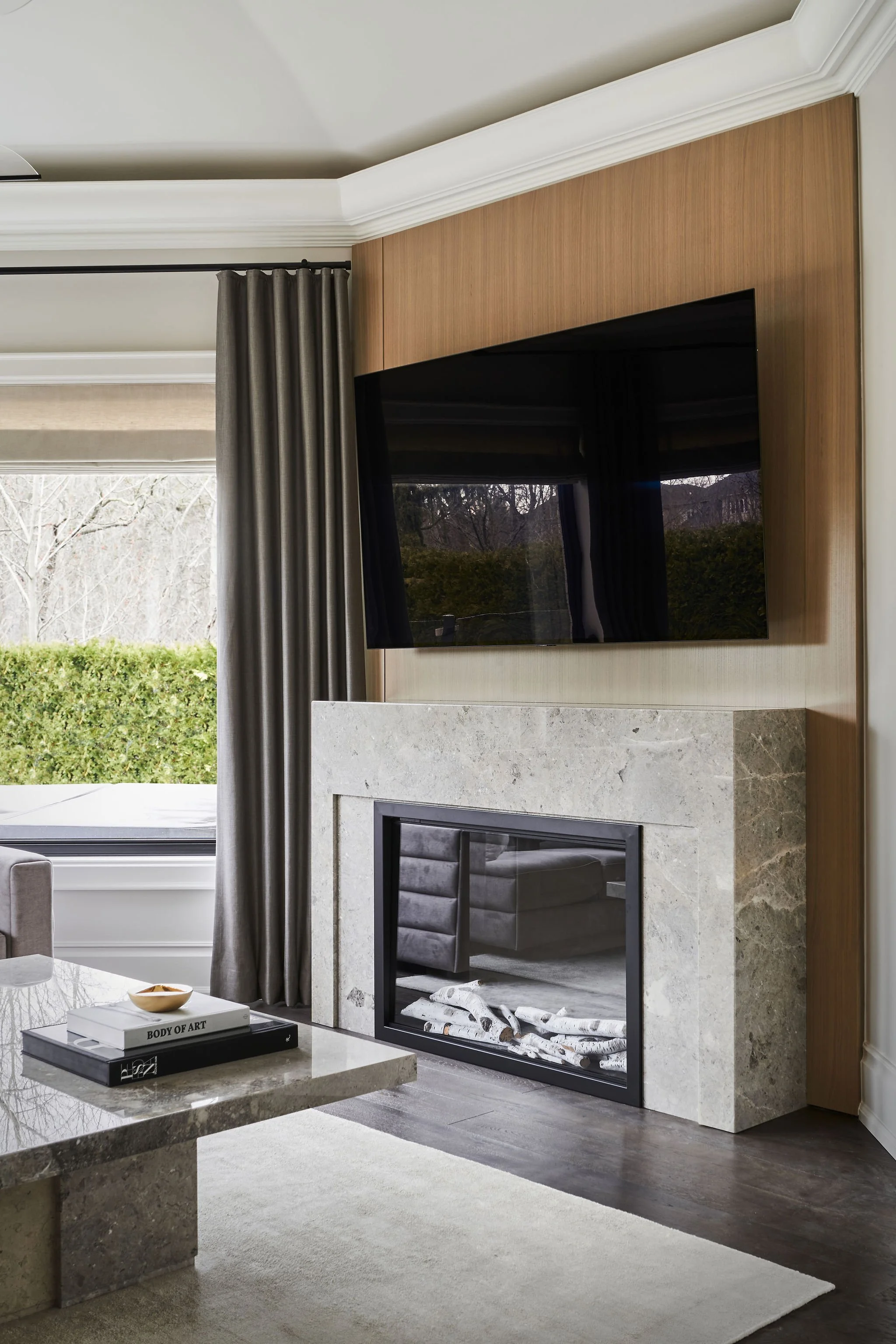 Living room with a fireplace, mounted flat-screen TV, large window with grey curtains, coffee table with books, and a white area rug.