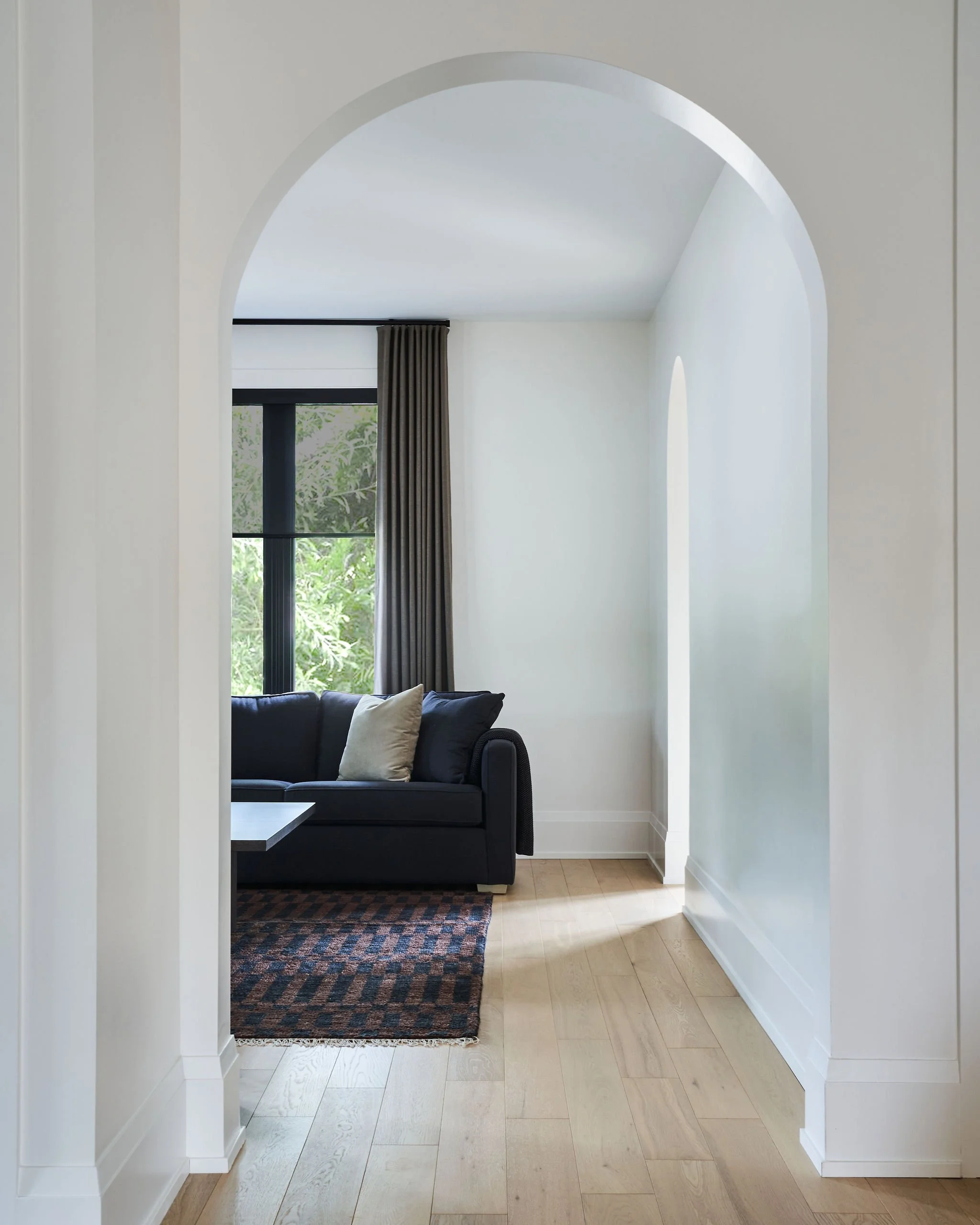 View of a living room seen through an arched doorway, with a black sofa, beige pillows, a window with dark curtains, and a patterned rug on light wooden floors.