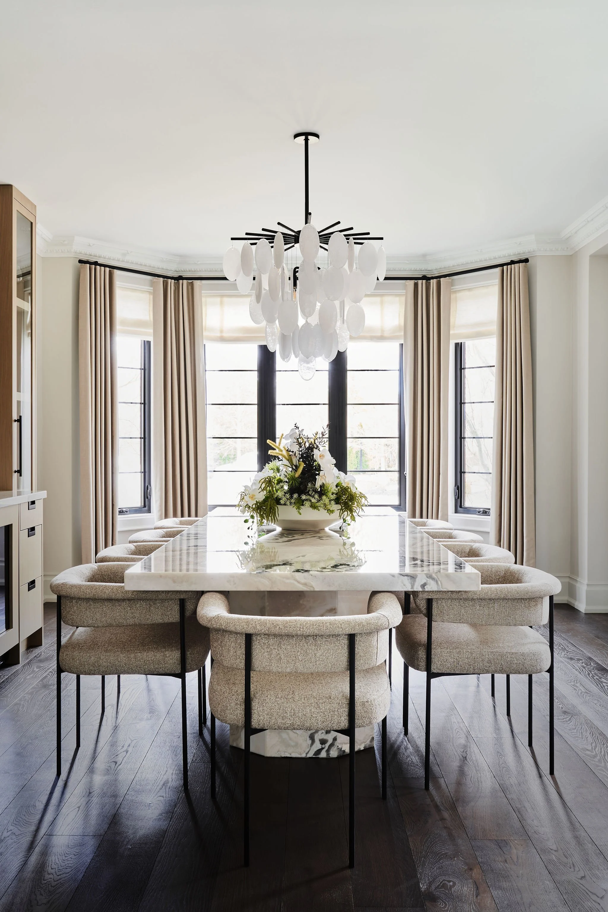A dining room with a marble-topped table surrounded by beige upholstered chairs with black metal legs, a modern chandelier hanging from the ceiling, large windows with beige curtains, and a floral centerpiece on the table.