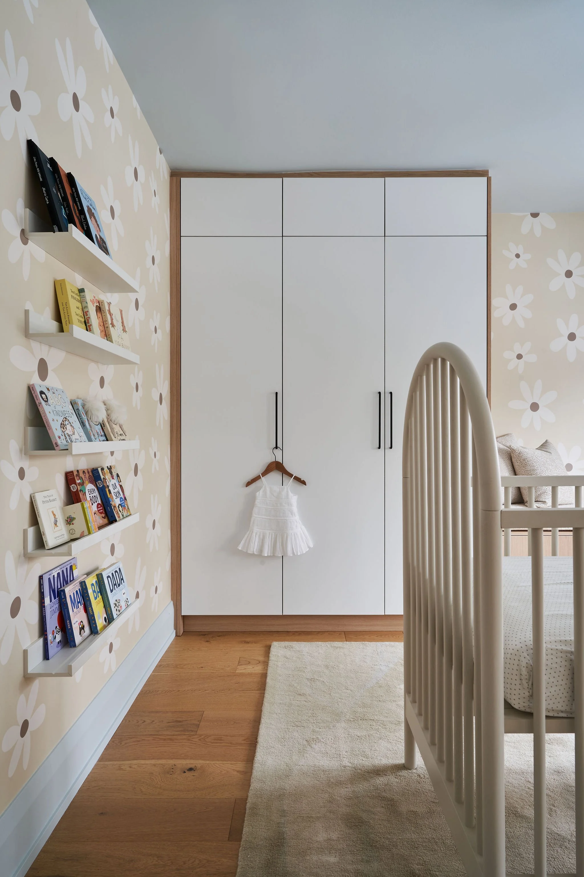 A cozy children's bedroom with floral patterned wallpaper, white bookshelves filled with books, a large white wardrobe with black handles, a beige crib with a white mattress, and a cream rug on wooden flooring.