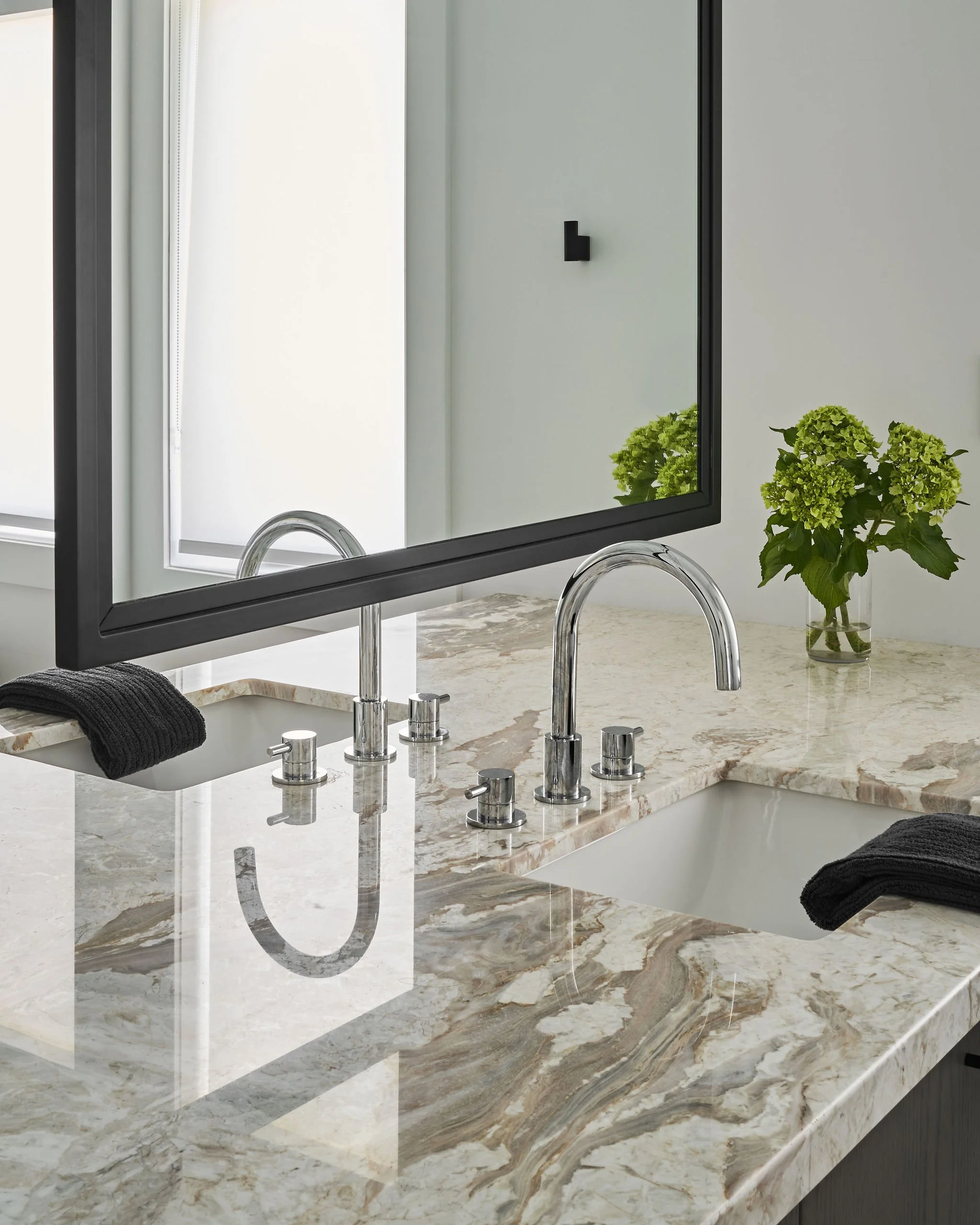 Close-up of a bathroom counter with a marble surface, a sink, and a large mirror. There are two black towels on the counter and green hydrangea flowers in a glass vase. The mirror reflects a portion of the plain wall with a small black object and a w