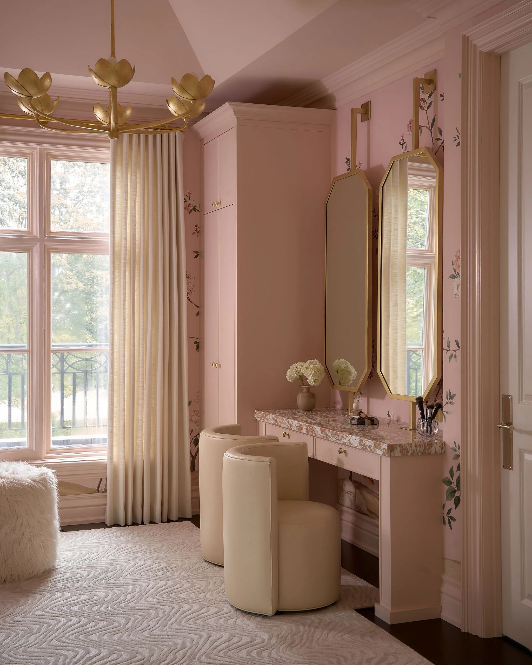 A corner of a pink cottage-style bedroom with three windows and a vanity with a marble top, two white upholstered chairs, a mirrored wall, floral wallpaper, a golden chandelier, and a fluffy white rug.
