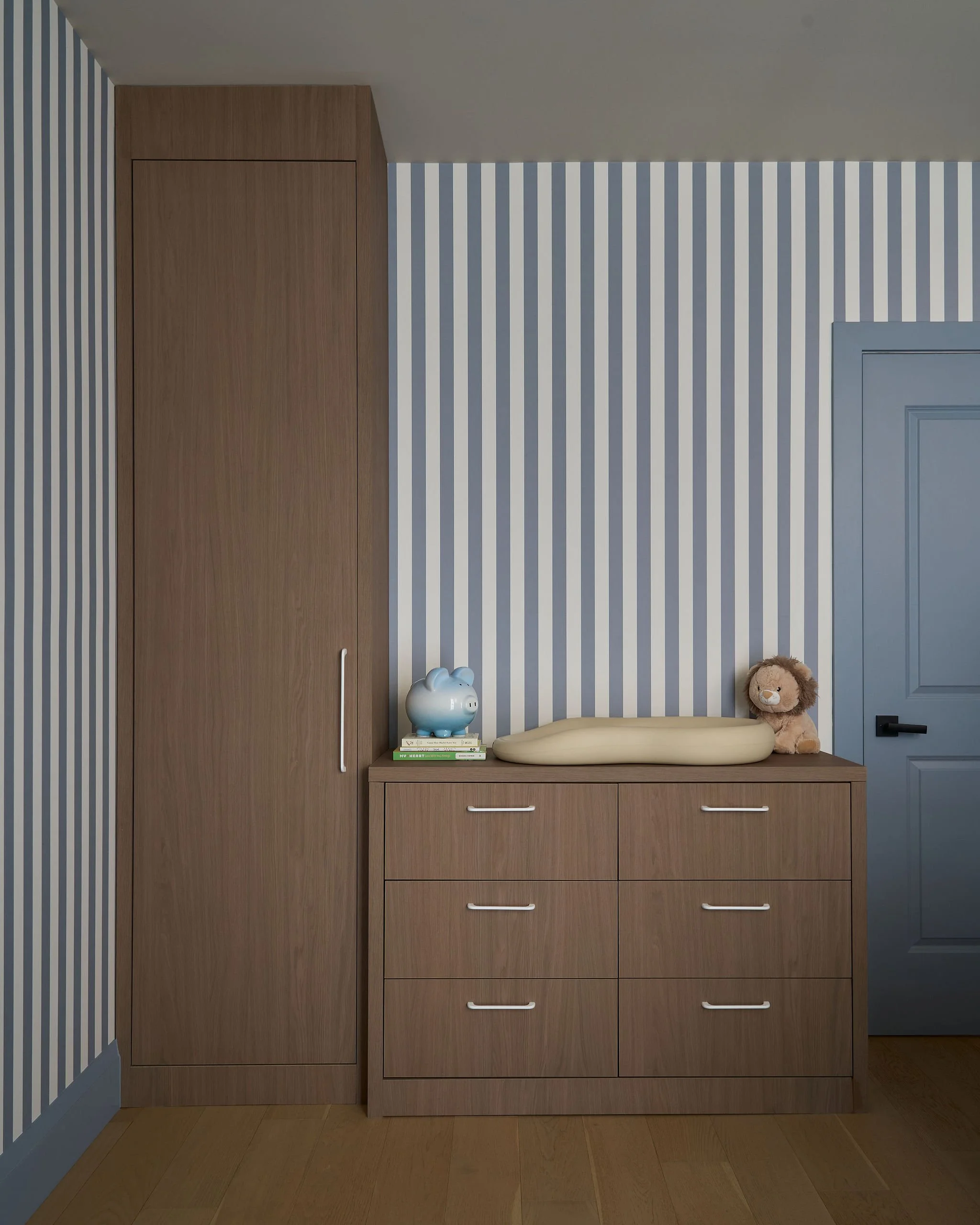 A wooden built-in closet with a vertical handle on the left side, a dresser with four drawers and white handles below a shelf, a piggy bank, a small stack of books, and a plush lion toy in a room with striped blue and white wallpaper and a blue door.