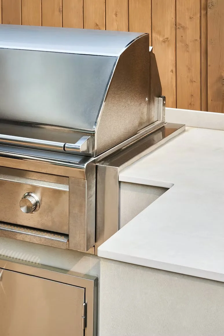 Stainless steel outdoor grill with a closed hood, positioned next to a white countertop and a wooden fence in the background.