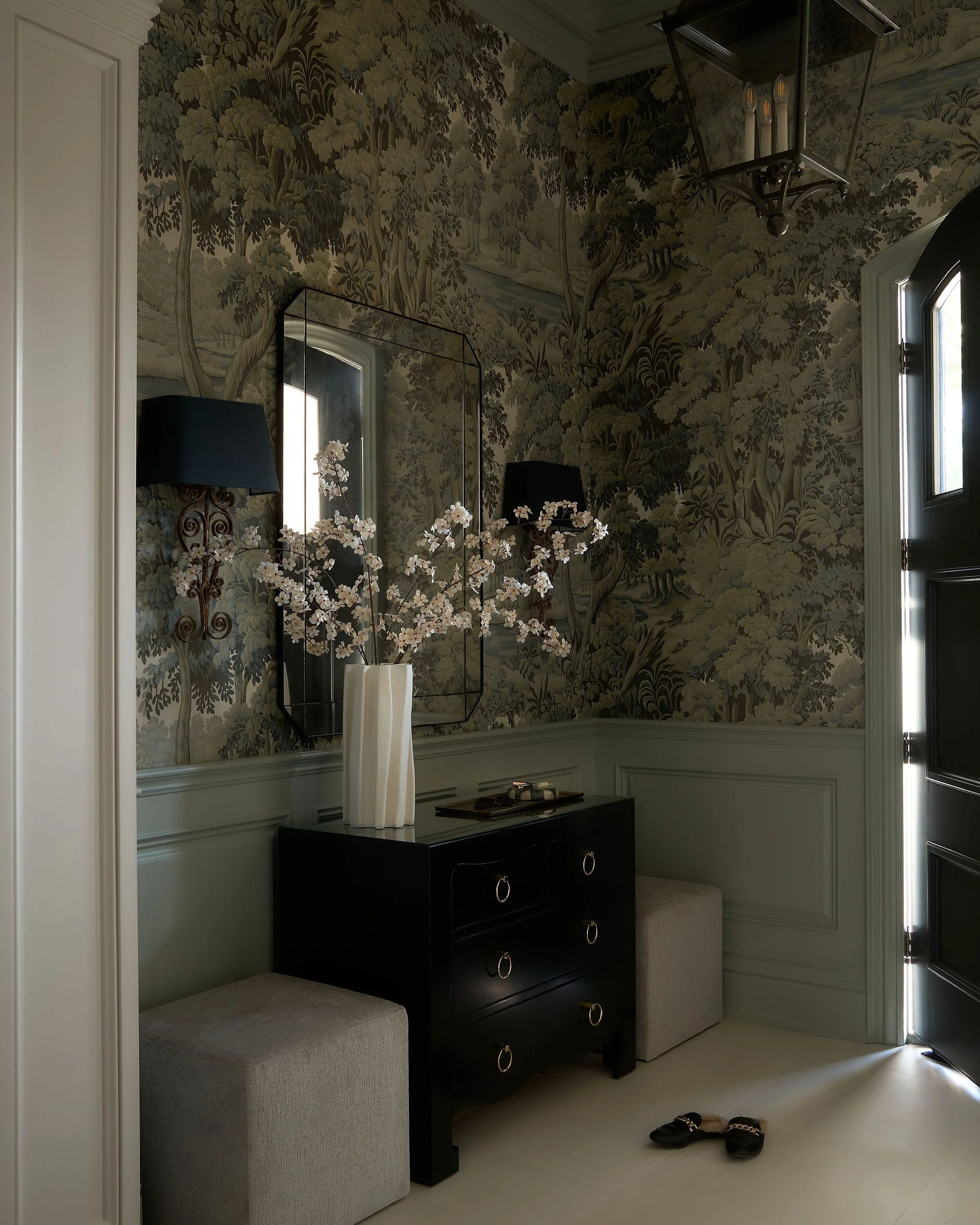 A corner of a room with dark furniture, a tall black chest with decorative rings, a beige cushioned bench, a mirror, a wall-mounted lamp with a black shade, a floral vase, and a wallpapered wall with a toile pattern depicting trees and nature scenes,