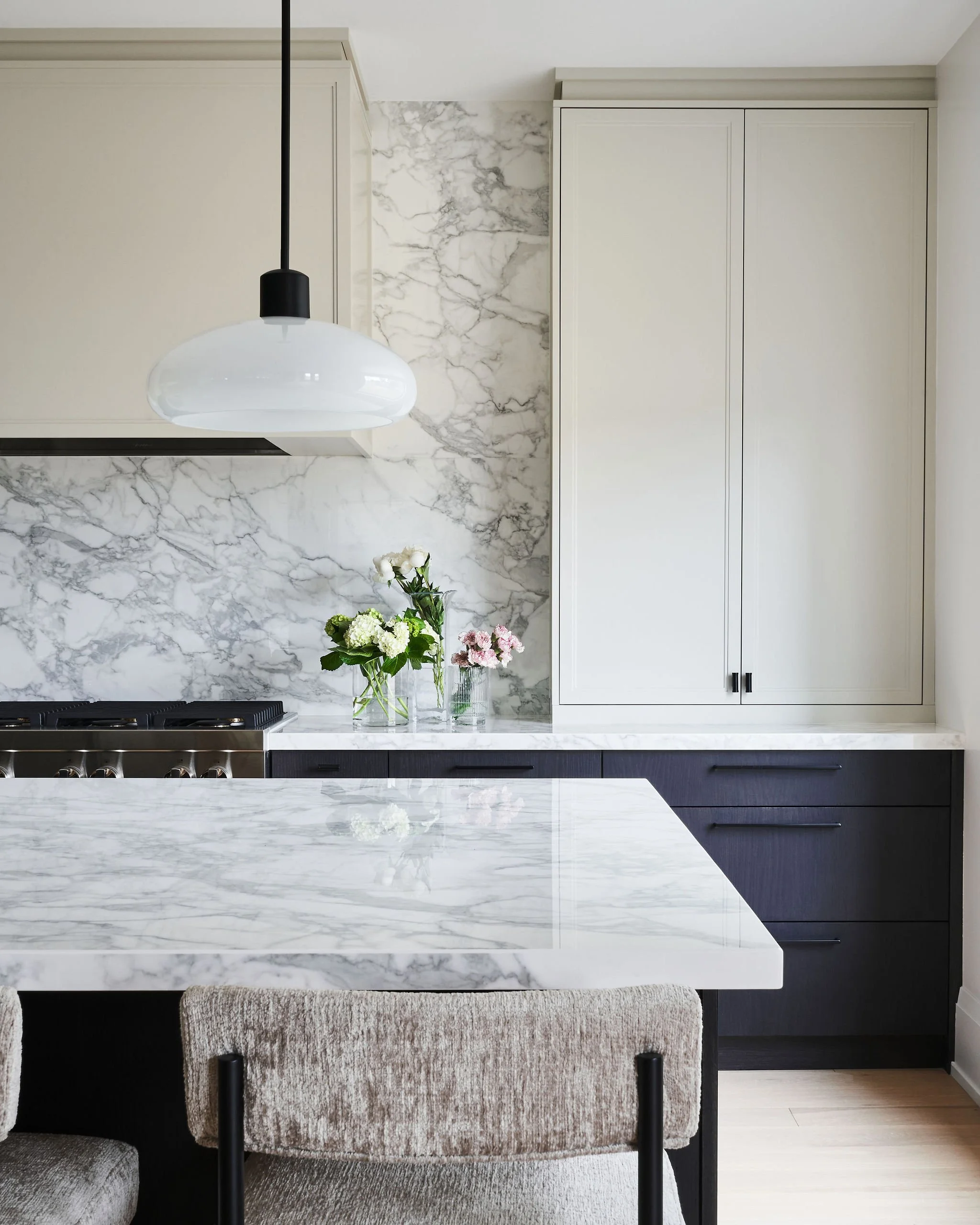A modern kitchen with a white marble island, a black and white pendant light, white cabinetry, floral arrangements on the counter, and light-colored flooring.