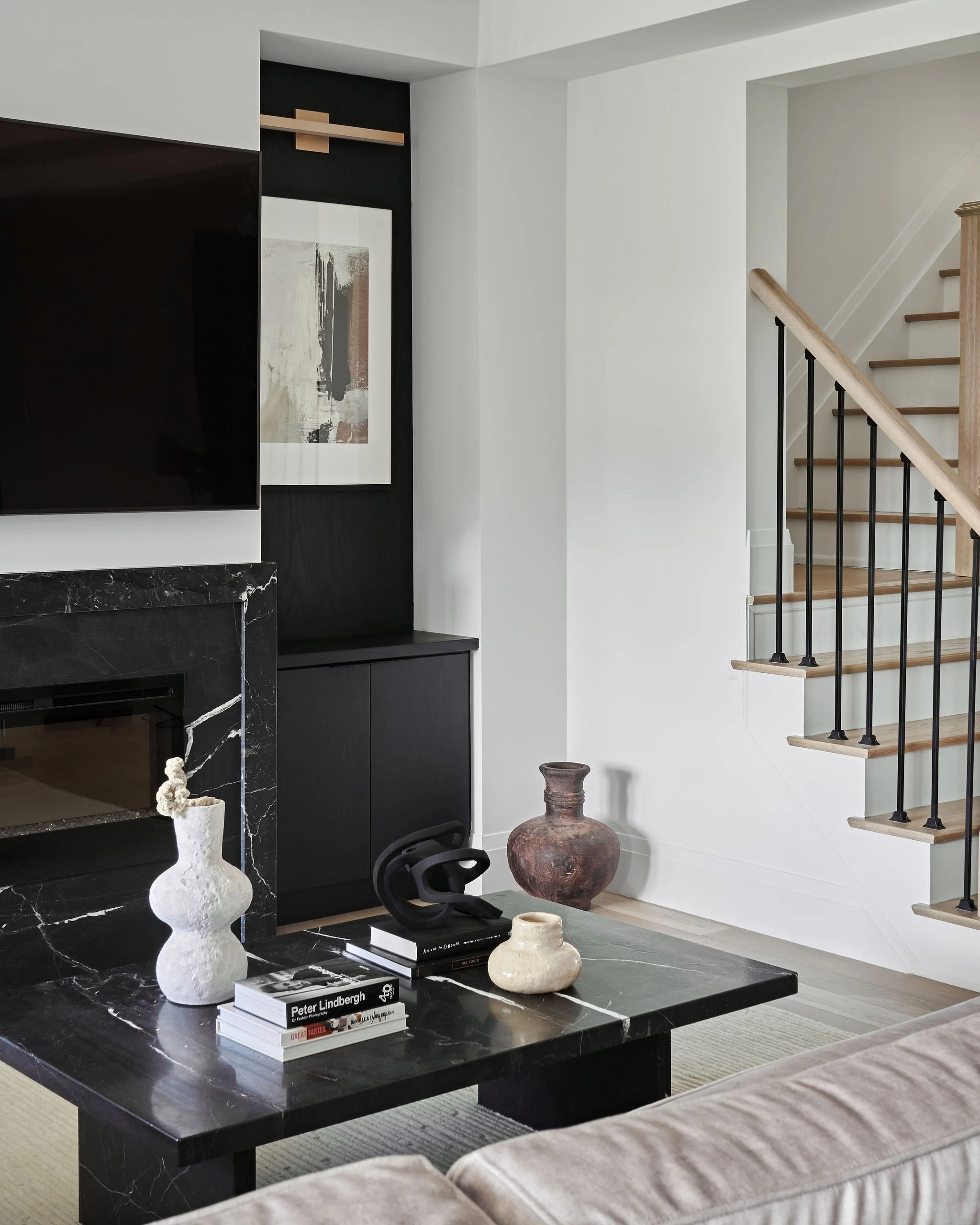 Living room interior with a black marble fireplace, a black coffee table with books and decorative vases, a large abstract artwork on a black wall, a staircase with wooden steps and black metal railing, light-colored sofas, rug, and decorative object