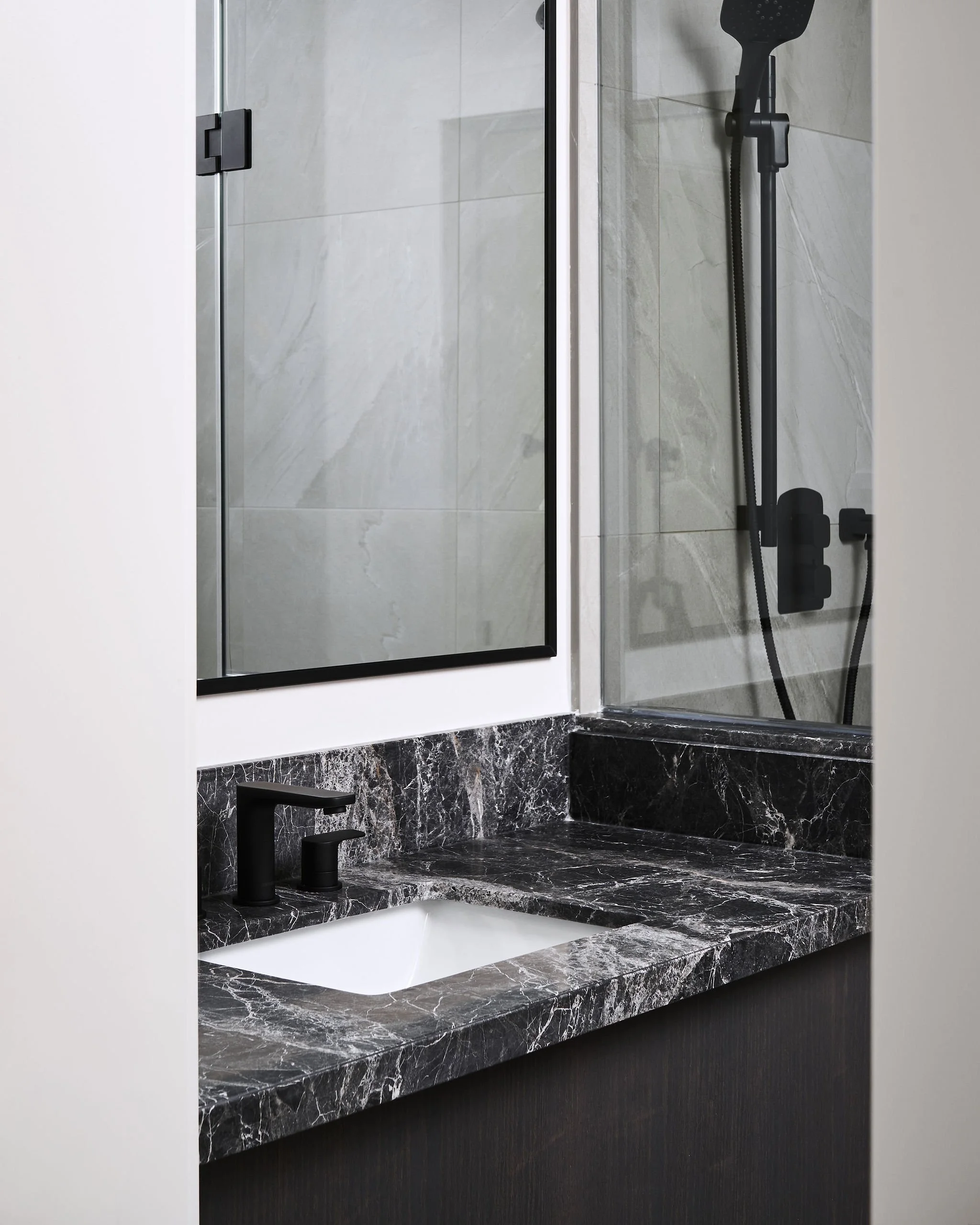 A modern bathroom vanity with a black marble countertop, integrated white sink, black matte faucet, and a large mirror. The mirror reflects a walk-in shower with glass door, gray tiled walls, and a black shower fixture.