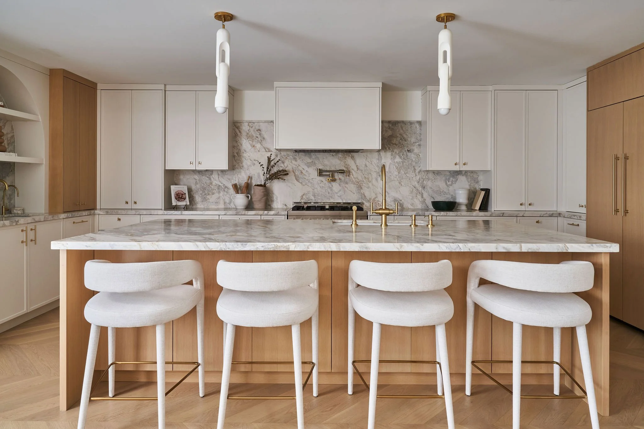 Modern kitchen with white marble island and matching backsplash, gold fixtures, white and wood cabinets, decorative items, and four white upholstered chairs.