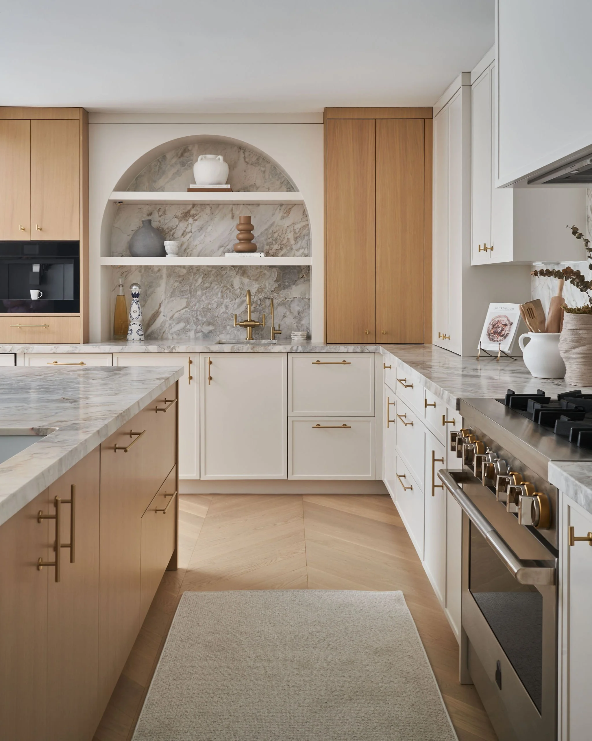 Modern kitchen with white and wood cabinets, marble countertops, gold hardware, and built-in appliances, including an oven and gas stove. A white rug is on the light wood floor.