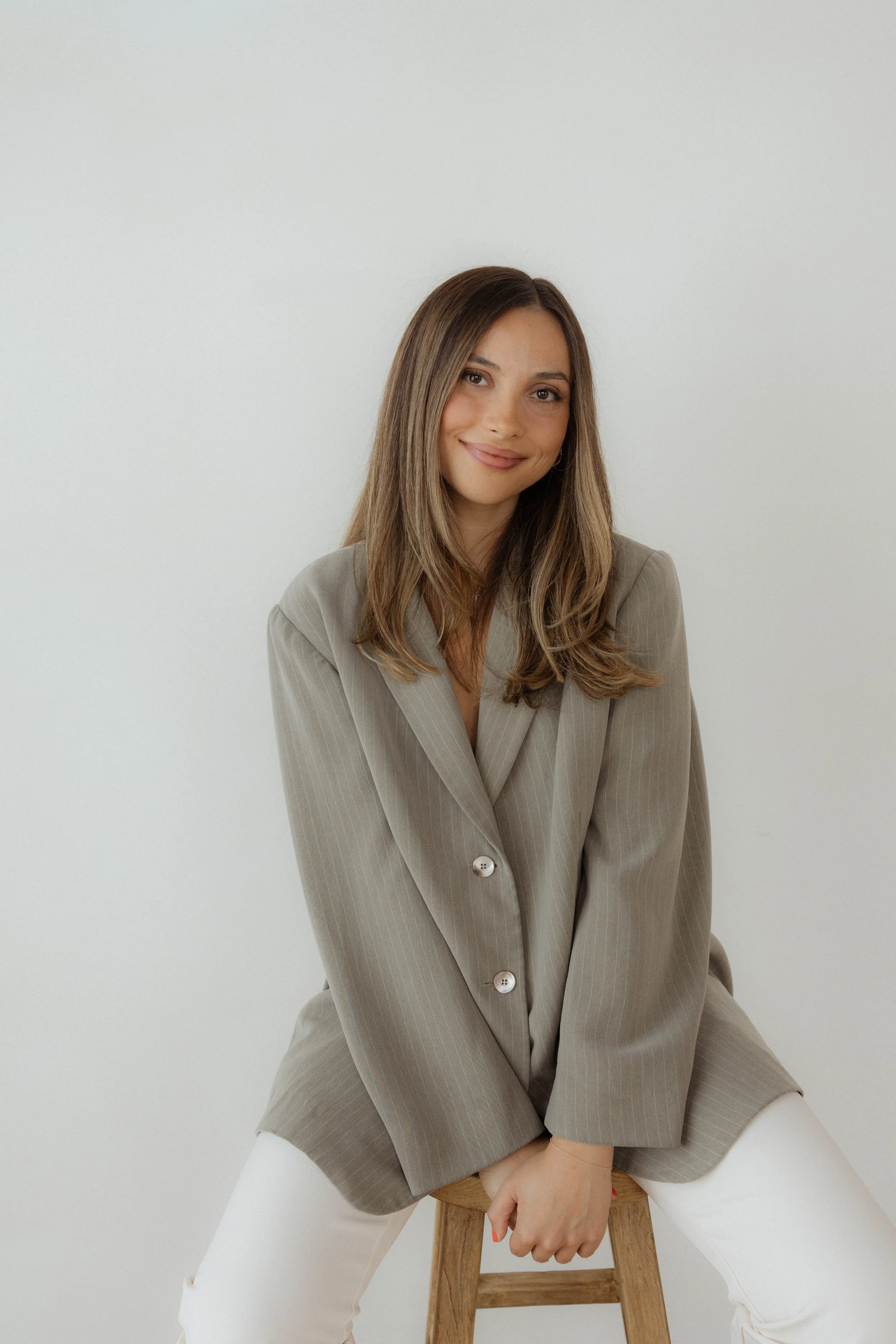 A woman sitting on a wooden stool, smiling, wearing a beige blazer and white pants, with her hand resting on her knee against a plain white wall.