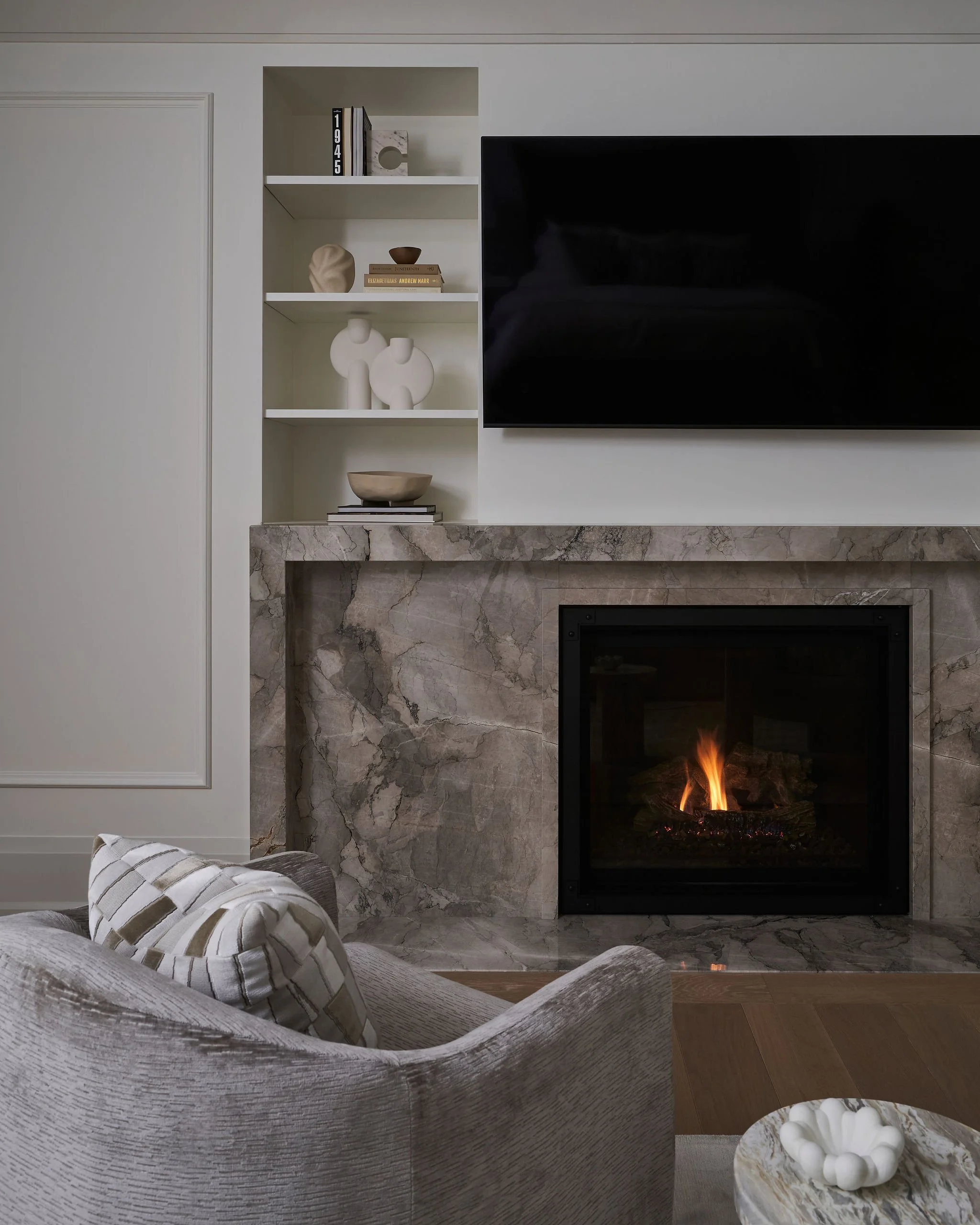 Living room with lit fireplace, white built-in shelf with decorative arts including vases and books, a mounted large flat-screen TV, and a marble surface with a decorative bowl filled with white shells.