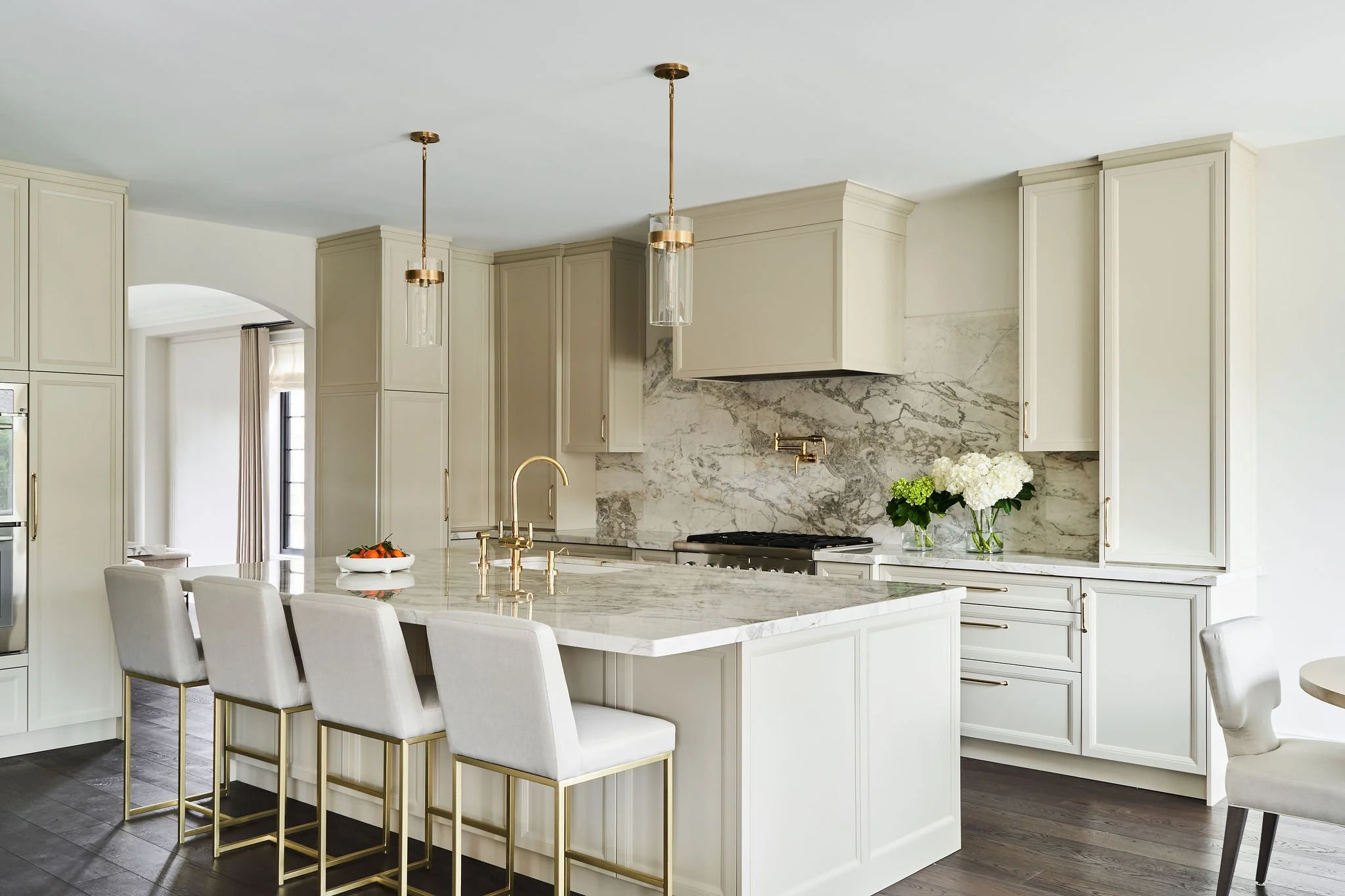 Modern white kitchen with marble backsplash, gold fixtures, and an island with dining chairs.