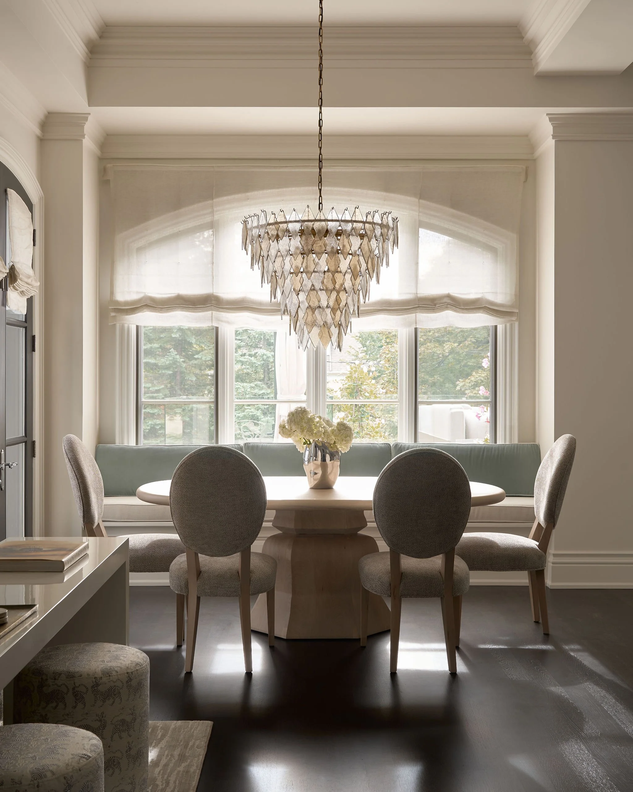 Dining area with a round table, six upholstered chairs, a large window with sheer curtains, a floral centerpiece, a chandelier, and sunlight streaming in.