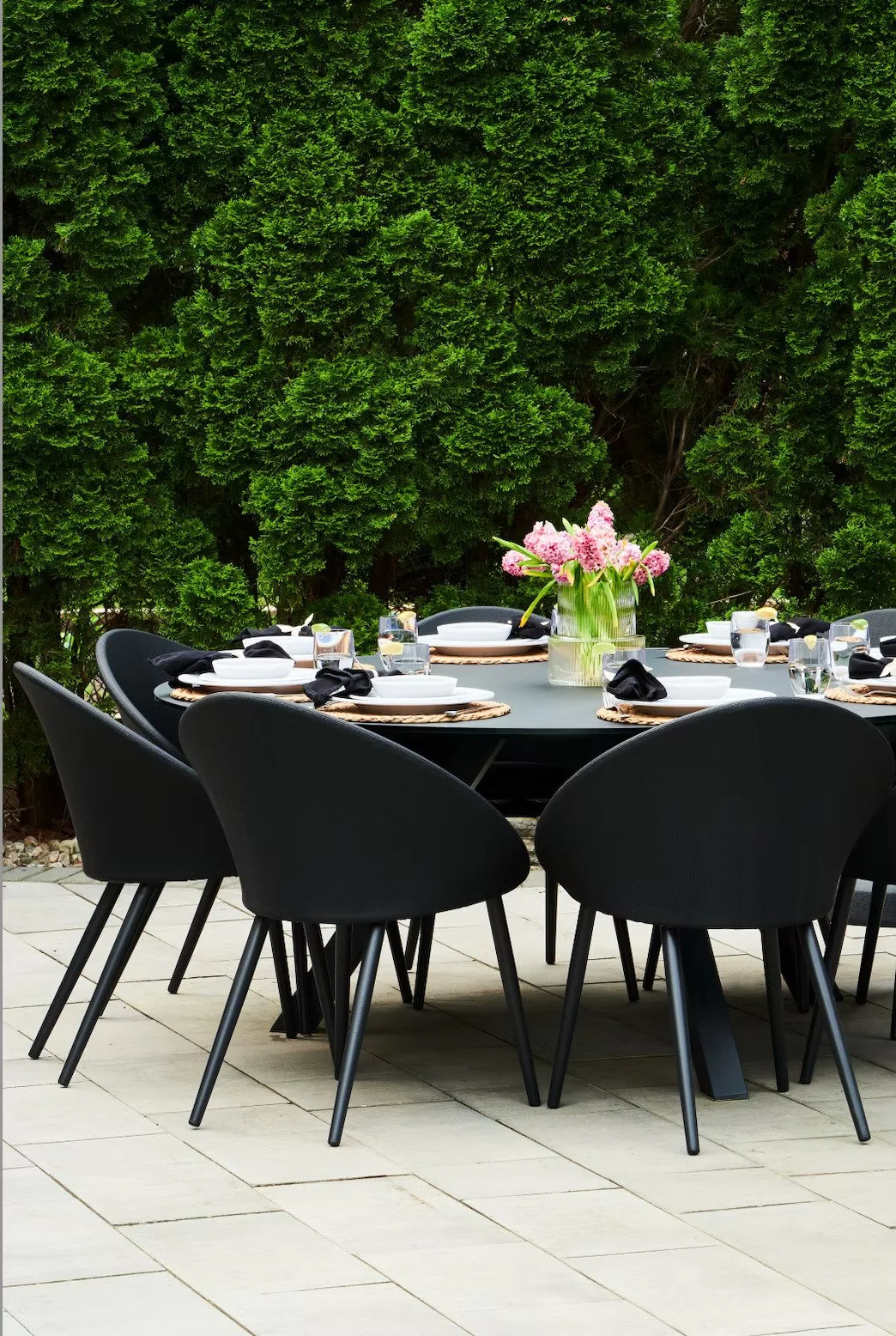 Outdoor dining table set with black chairs, a vase of pink flowers, white plates, black napkins, and glasses, surrounded by tall green bushes.