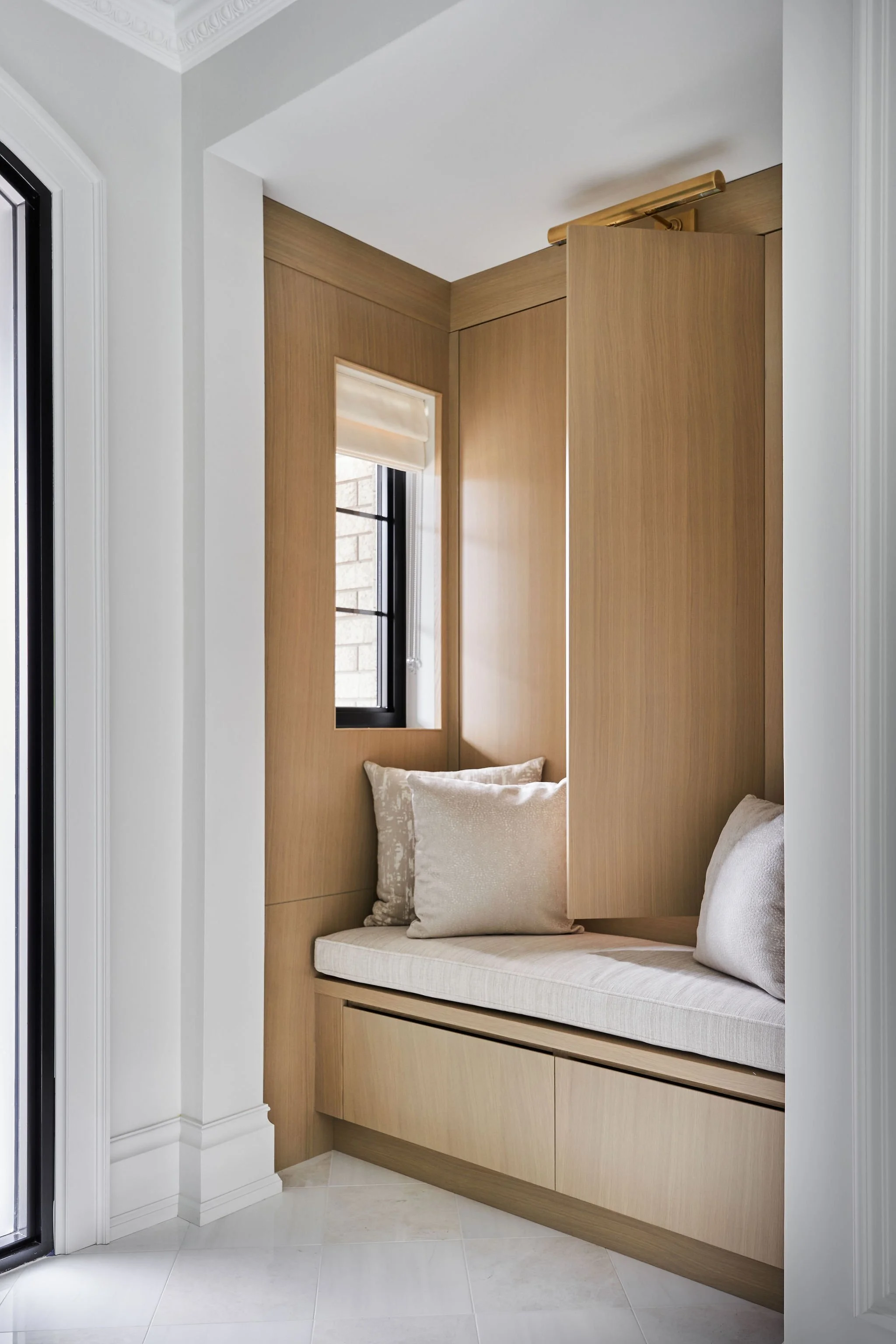 Built-in window seat with beige cushions and pillows, surrounded by wooden wall paneling, in a bright room with a window and white trim.