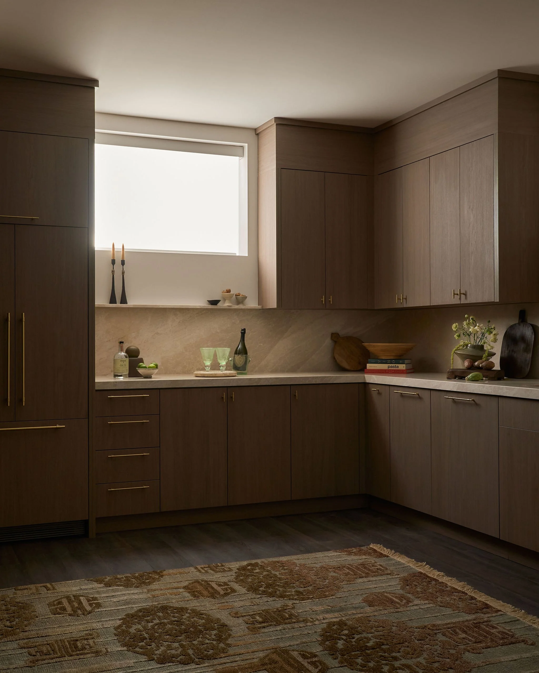 Modern kitchen with wooden cabinets, marble countertops, and a window with white blinds. Decor includes candles, bowls, wine bottles, books, and plants.