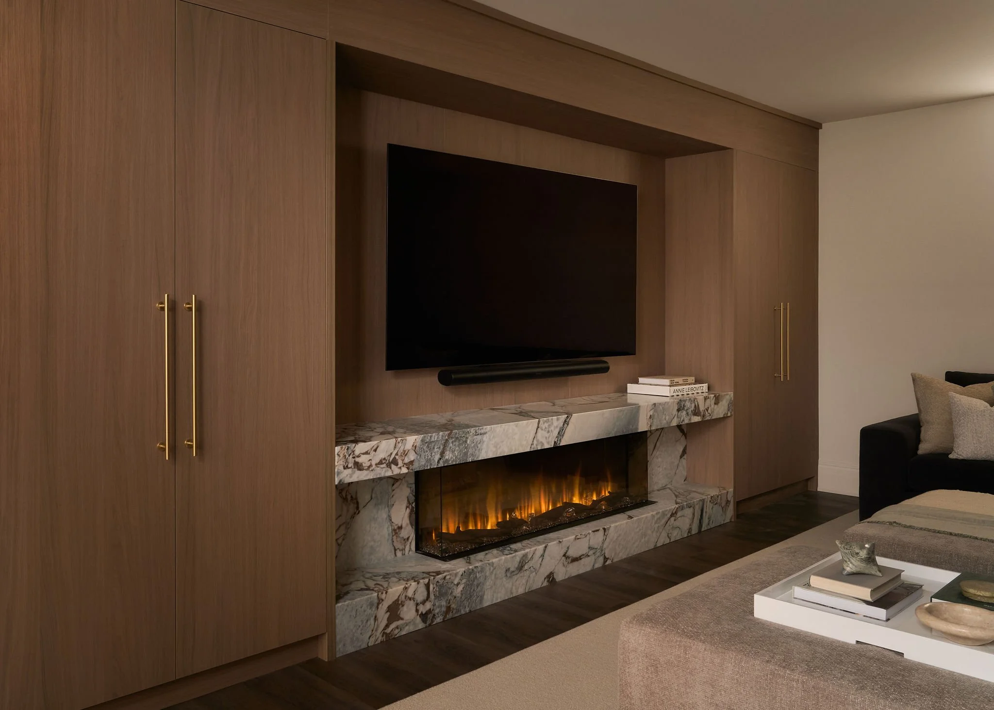 Living room with wooden cabinetry, a mounted flat-screen TV, a marble fireplace with a glowing fire, and a coffee table with books and decorative items.