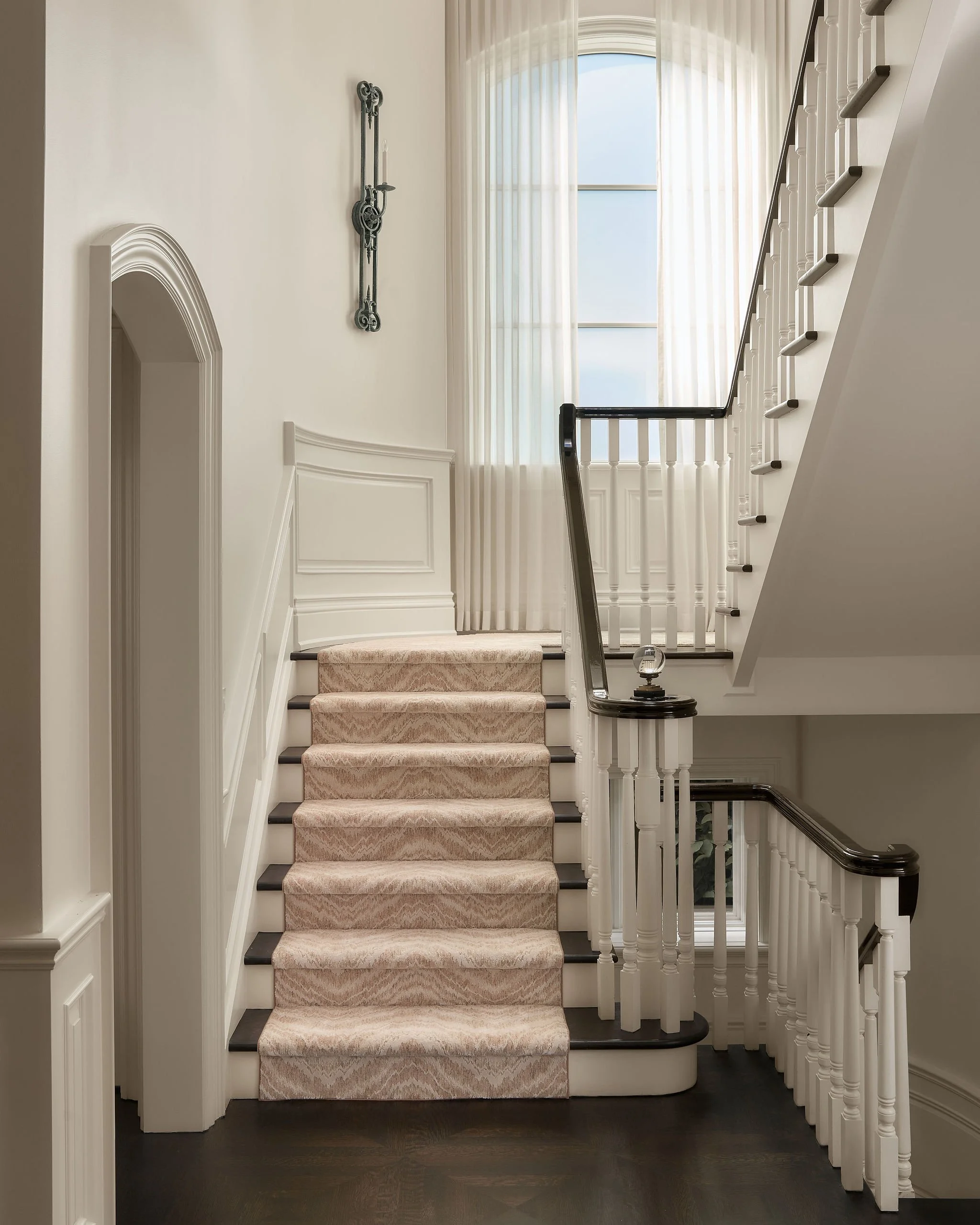 A staircase with beige carpeted steps, a dark wood handrail, white spindles, and a wall with decorative molding and a metal wall ornament. Large window with sheer curtains letting in natural light.