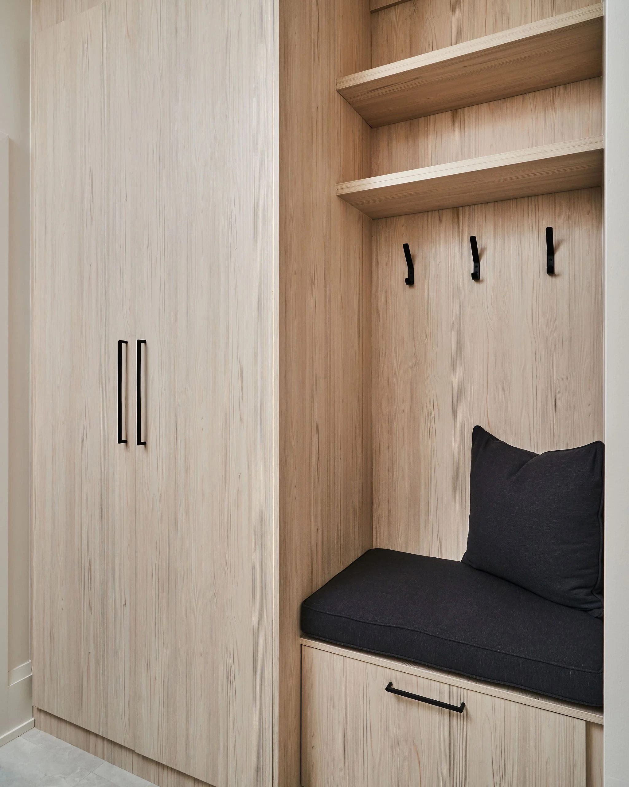 A built-in wooden closet with black handles, a black cushioned bench with a black pillow underneath, and black wall hooks on the pinkish wooden wall.