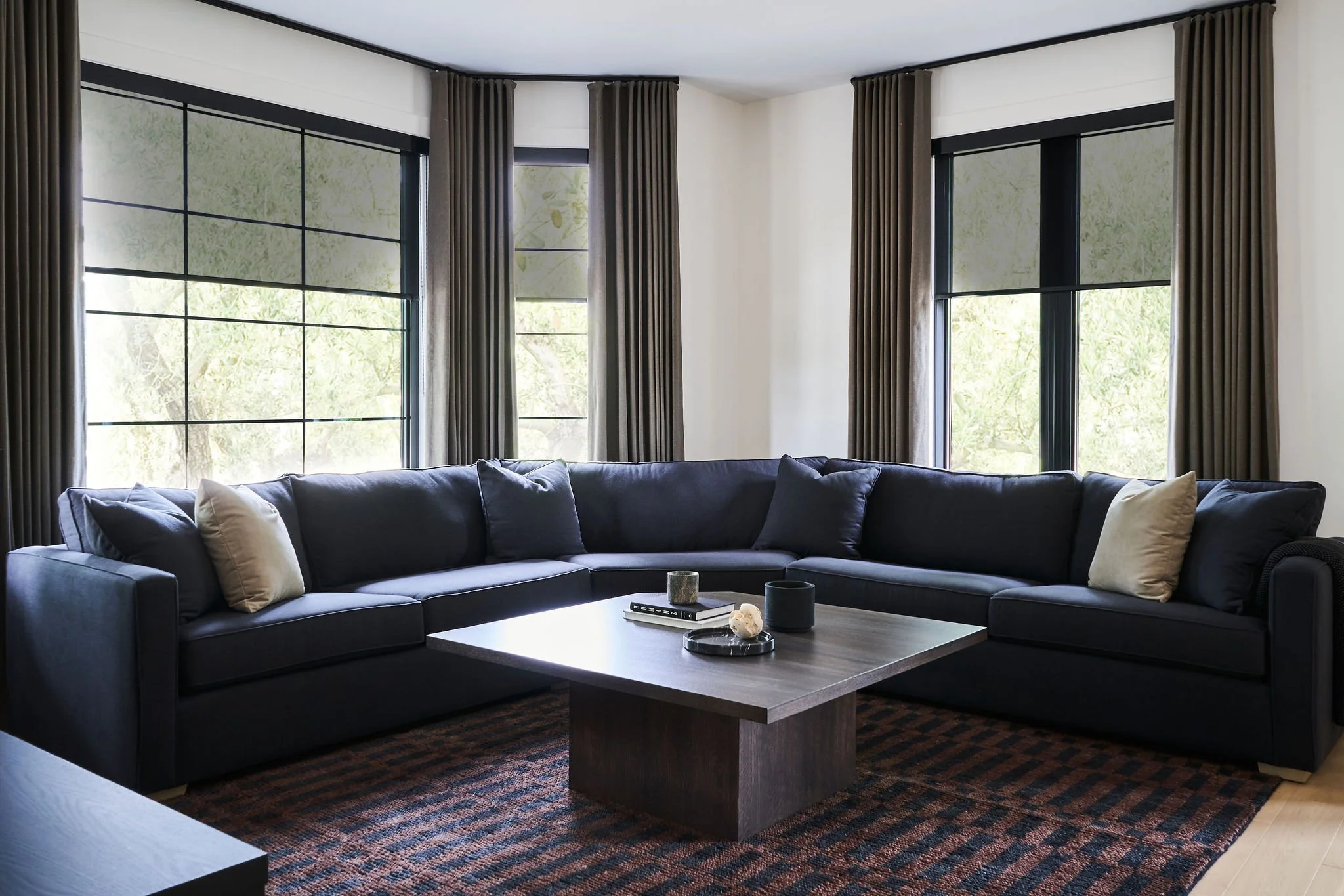Modern living room with a dark gray sectional sofa, beige pillows, a coffee table with decorative items, tall windows with blackout curtains, and a patterned rug.