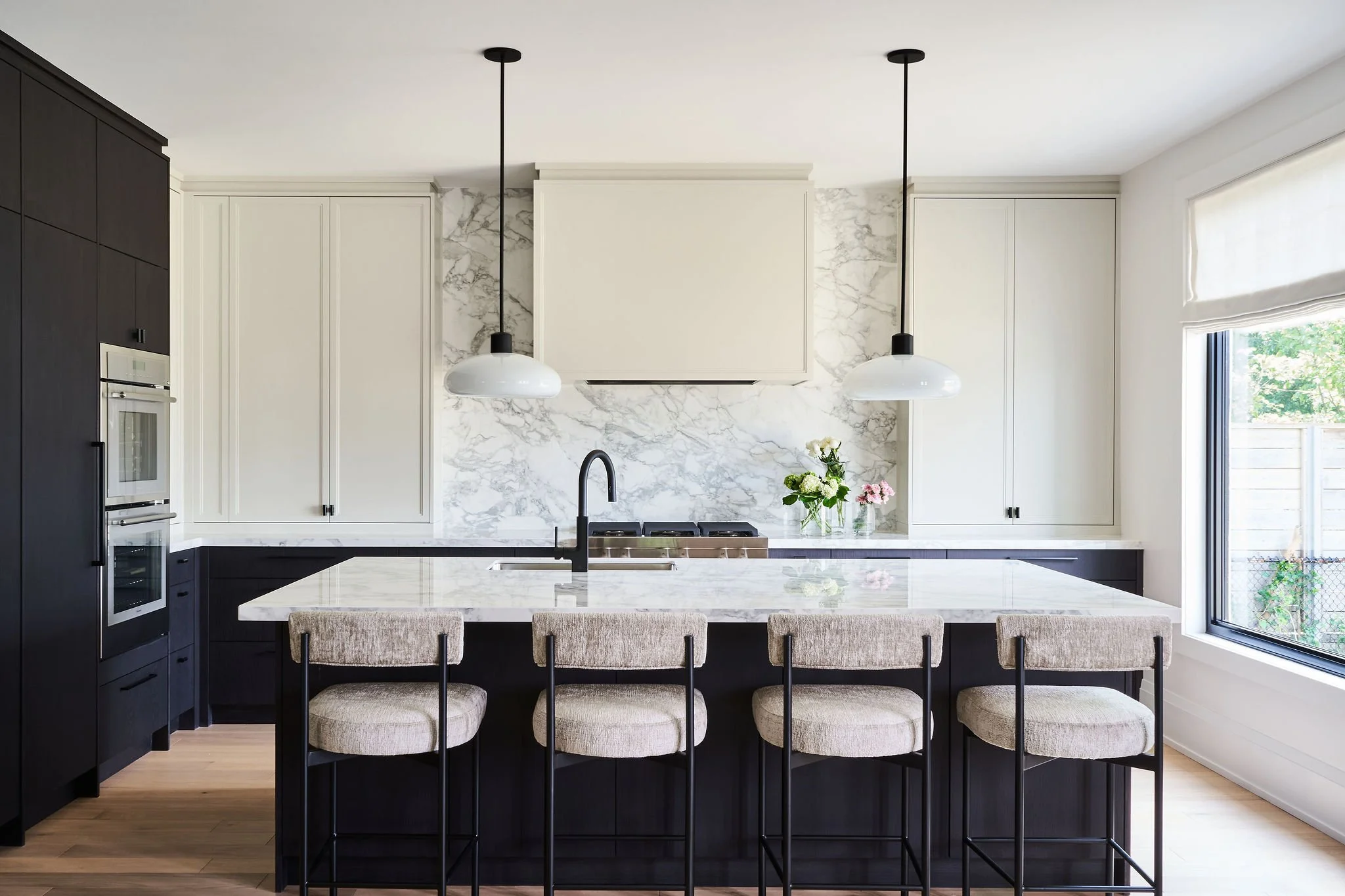 Modern kitchen with black and white cabinetry, marble backsplash, black pendant lights, large window, and a kitchen island with beige chairs and a vase of flowers.
