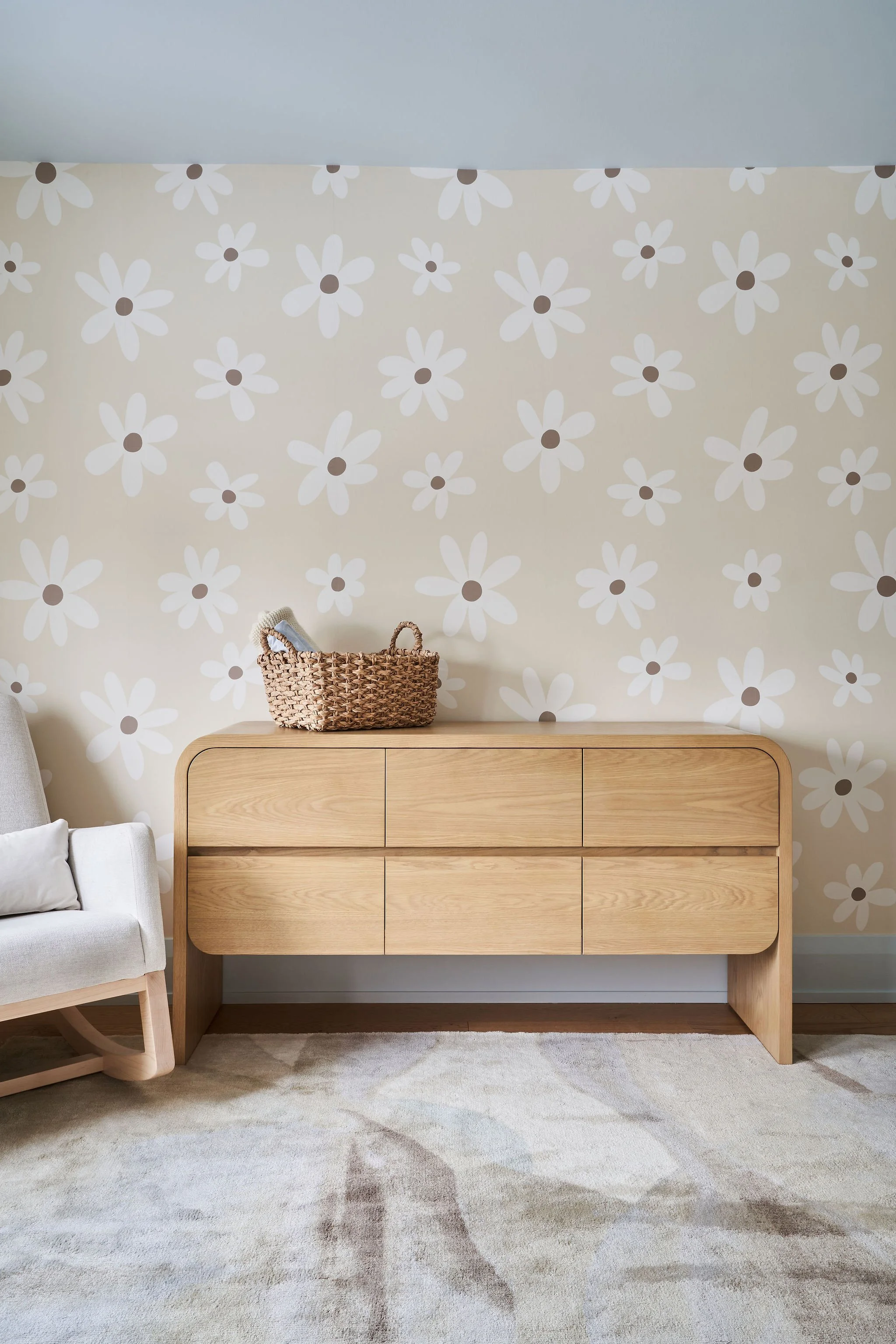 A wooden dresser with six drawers against a wall decorated with white daisy-patterned wallpaper. On top of the dresser is a woven basket with handles. To the left, part of a white armchair with a cushion is visible, and a patterned area rug covers th