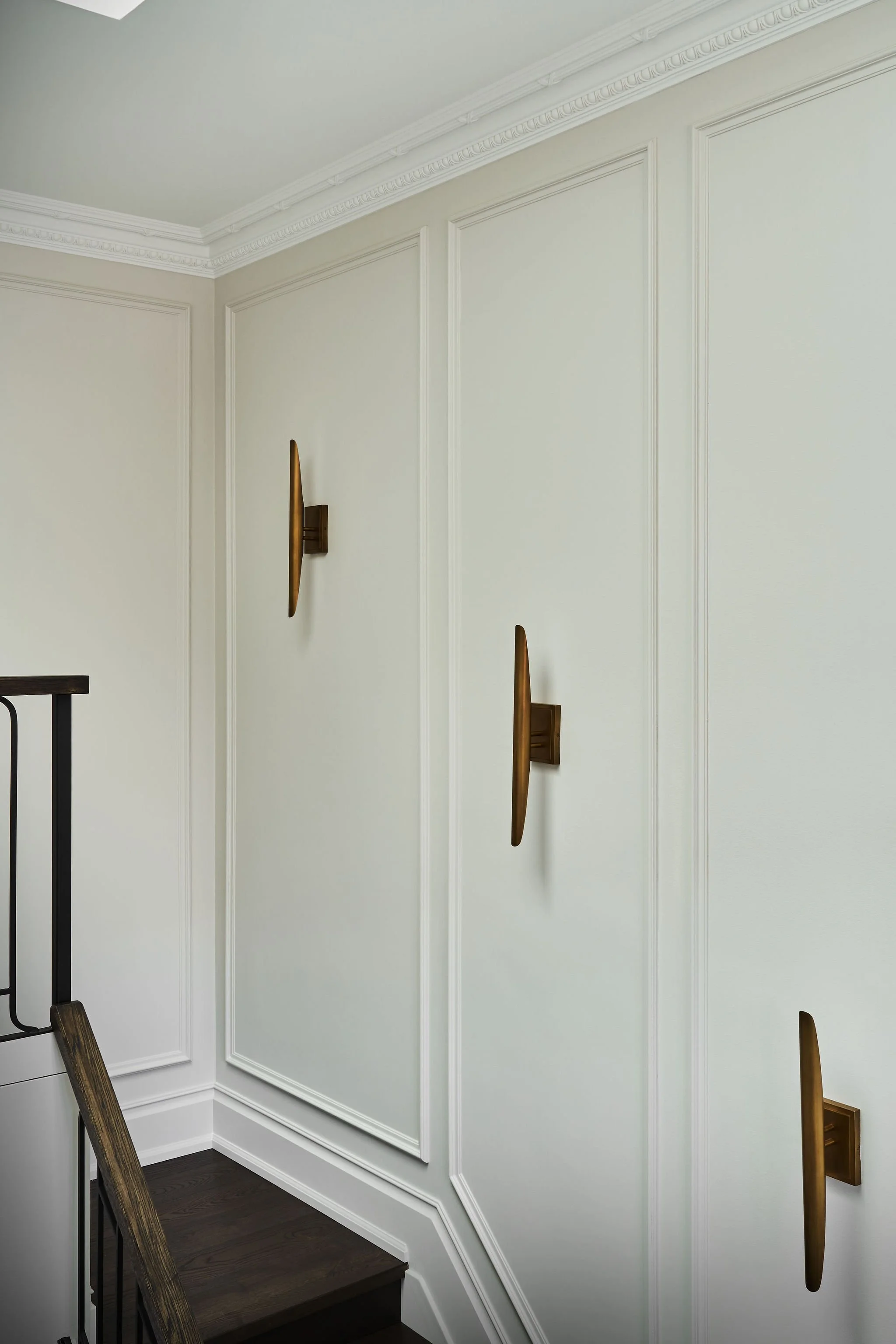 Inside a home, with light green walls with decorative molding, a staircase with wooden steps and a black metal railing on the left, and wall-mounted long, wood-colored art pieces.