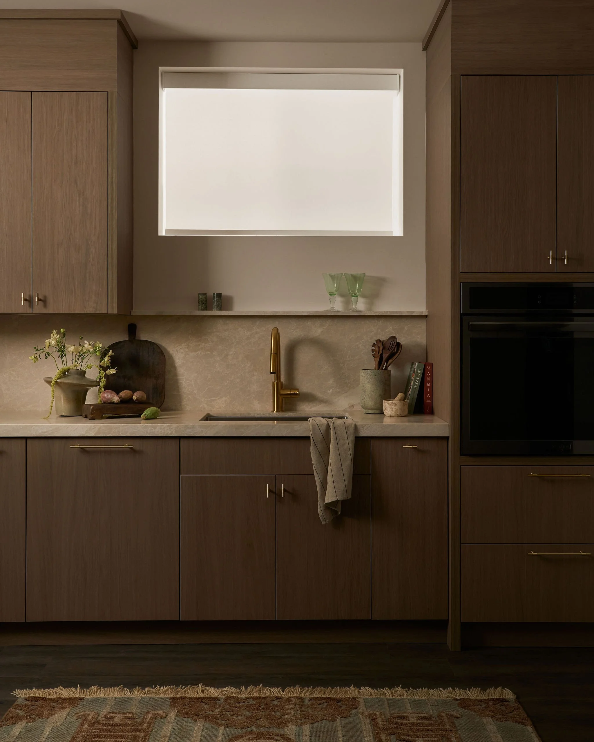 Kitchen with wooden cabinets, a window with closed blinds, a beige countertop, a gold faucet, decorative items including a green vase with flowers, a black cutting board, and books on the right side.