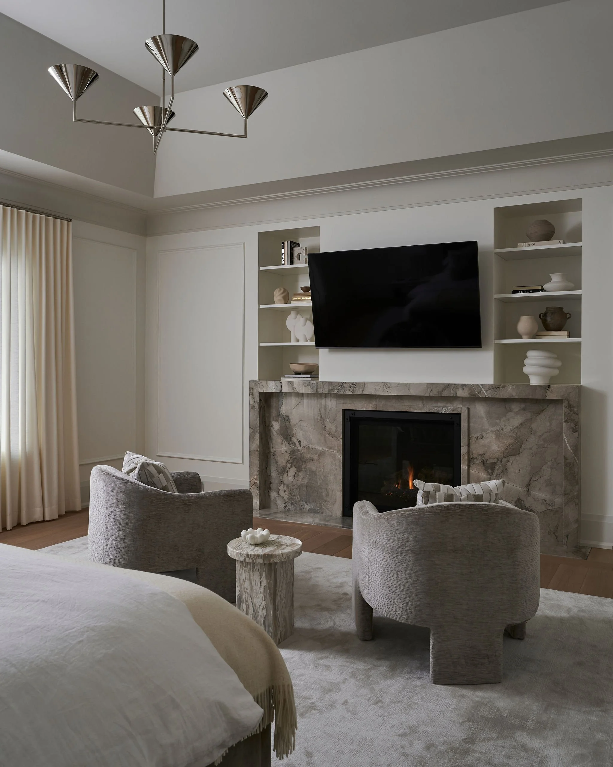 Living room with a marble fireplace, a wall-mounted TV, two beige upholstered armchairs, a small round marble side table with a white decorative piece, built-in shelves with decor and books, beige curtains, and a modern ceiling light fixture.