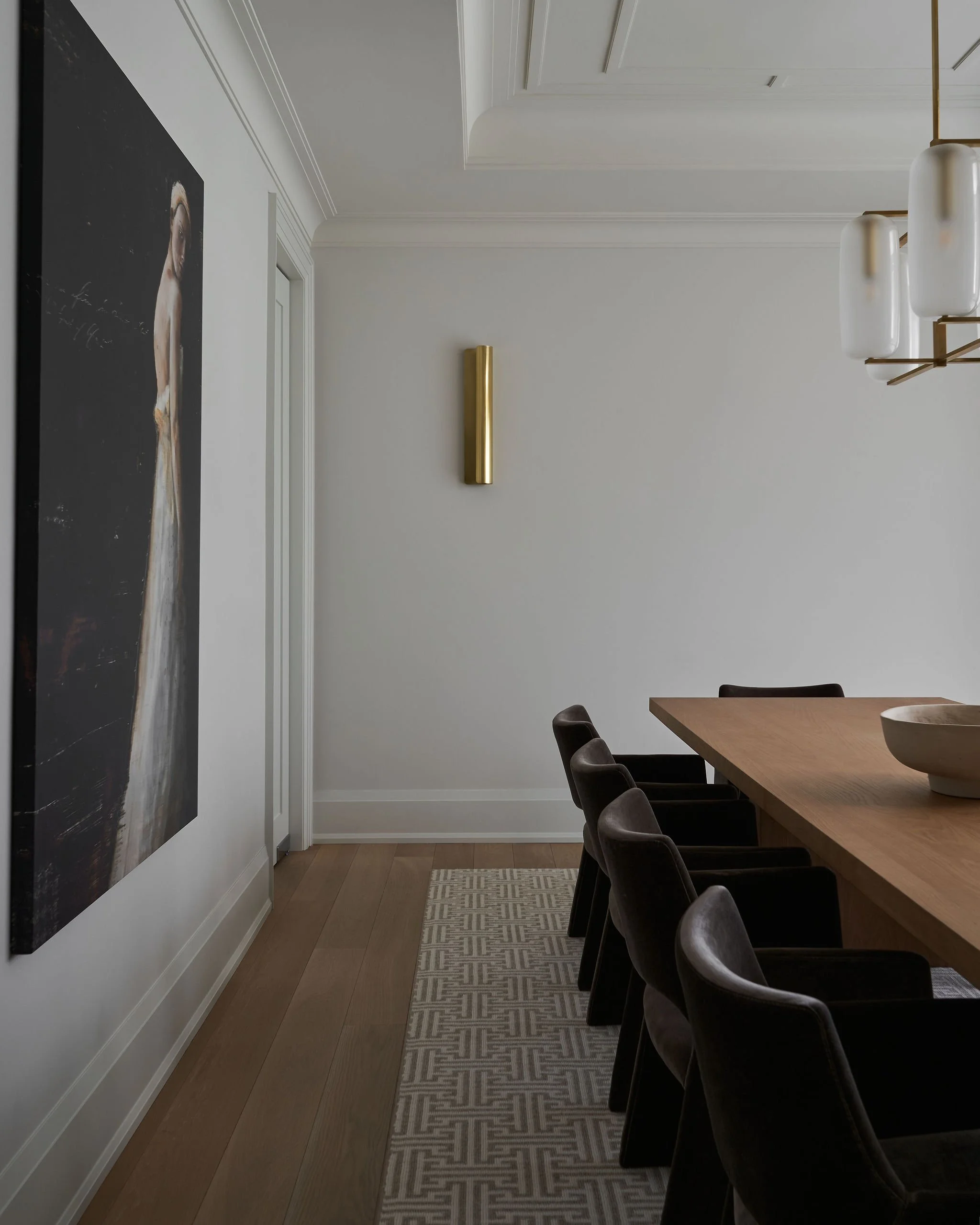 A modern dining room with a wooden table, black chairs, a white wall with a gold wall sconce, and a painting of a woman on the left side.