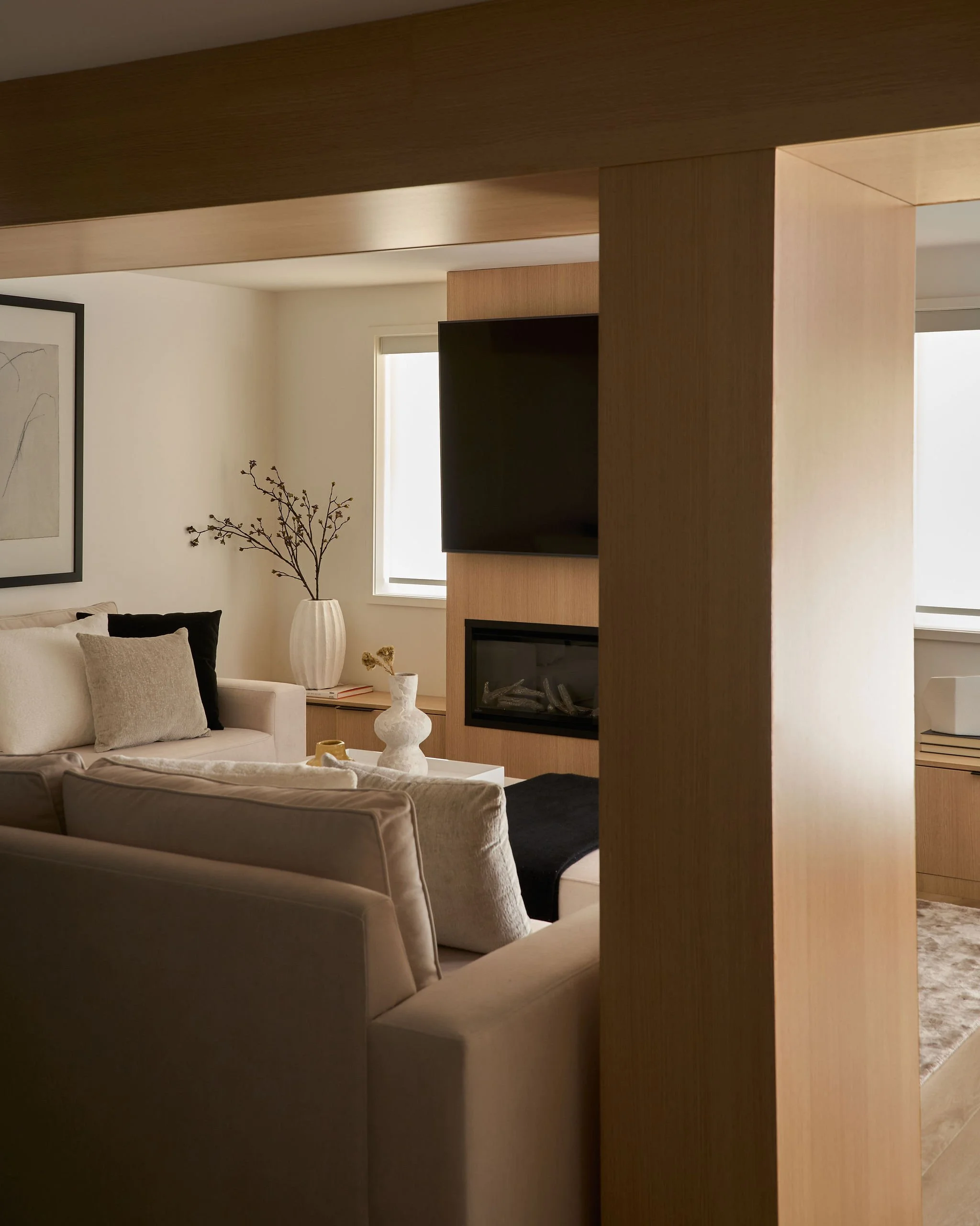 Living room with beige sofas, a white vase with a branch, a fireplace, a framed art piece on the wall, and windows bringing in natural light.
