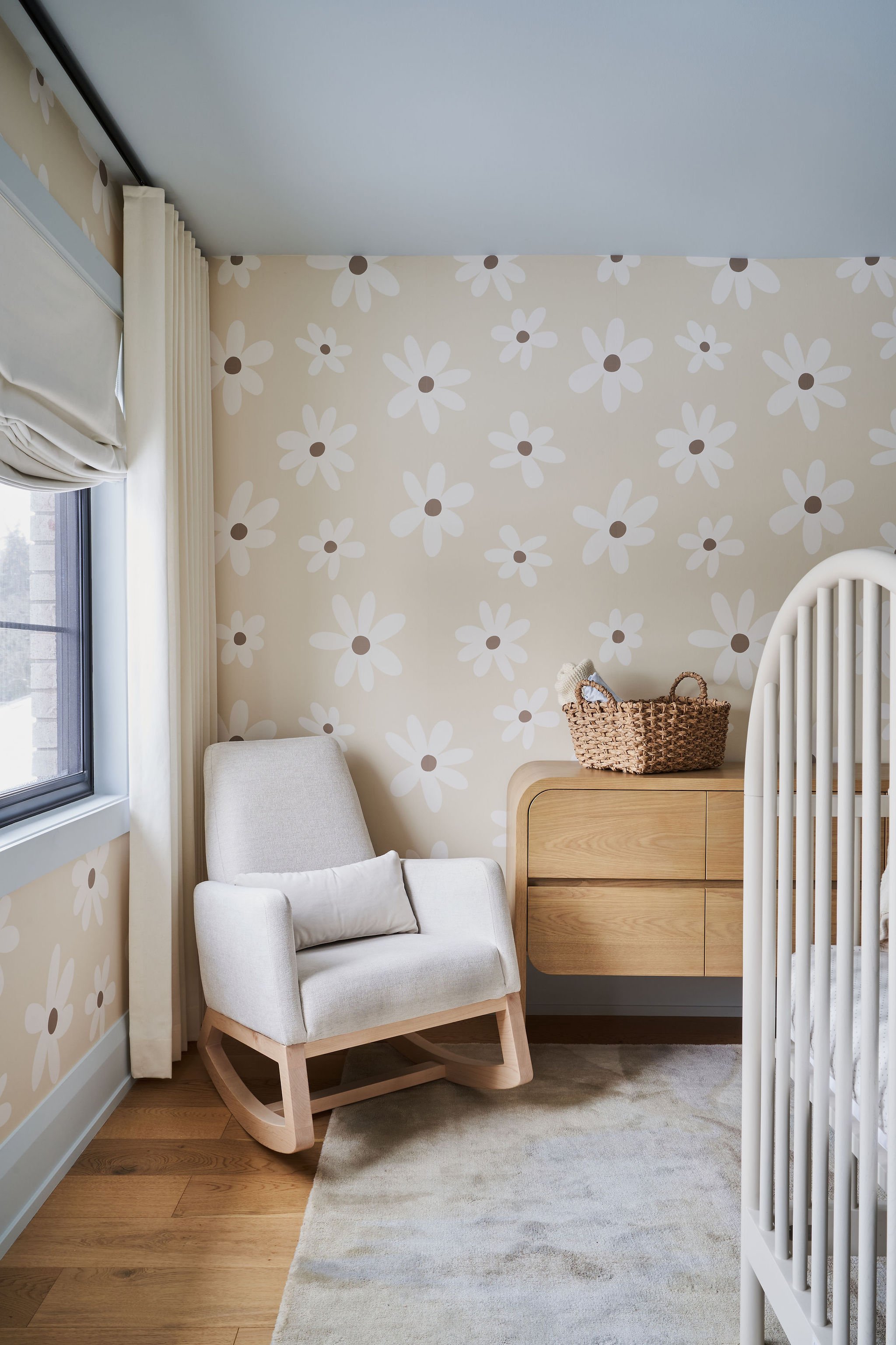 Cozy nursery with white rocking chair, beige floral wallpaper, wooden dresser, woven basket on top, window with white curtains, and beige carpeted floor.