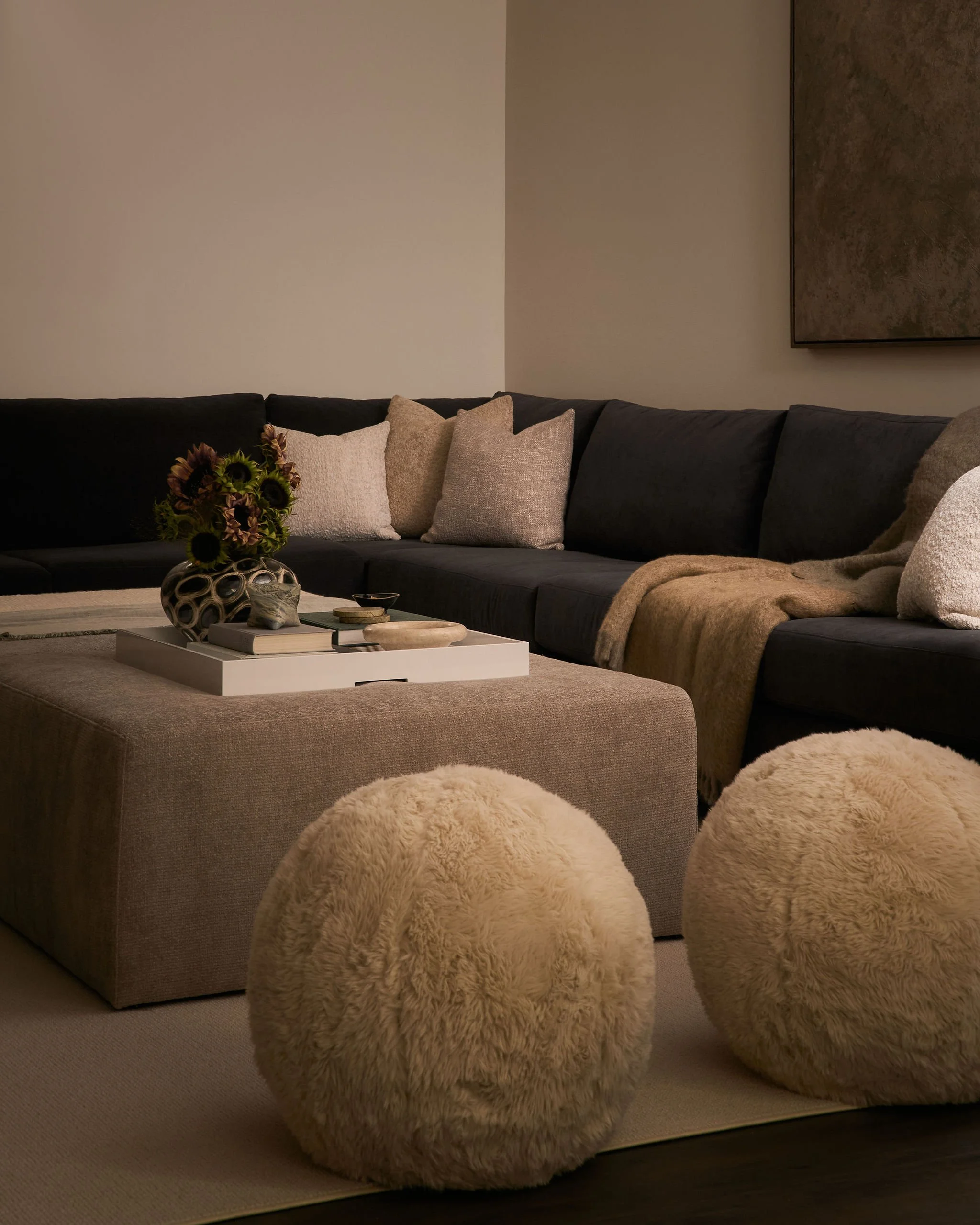 Living room with dark sofa, beige pillows, beige blanket, round plush ottomans, a white coffee table with books, a vase of sunflowers, and wall art.