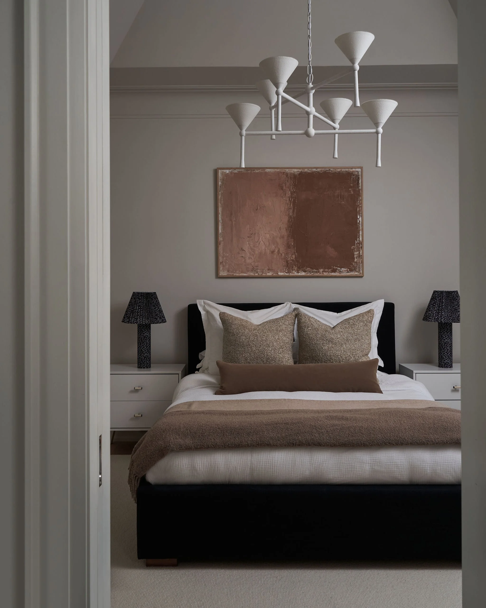 A bedroom with a bed, pillows, a brown blanket, nightstands with black lamps, and a modern chandelier hanging from the ceiling. There is an abstract artwork on the wall above the bed.