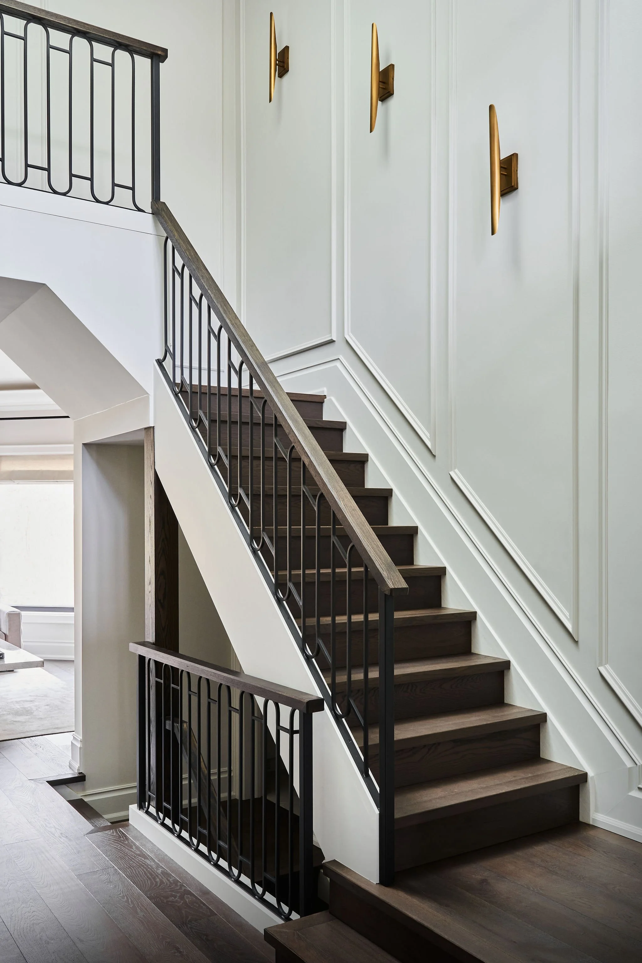 Interior staircase with dark wooden steps, white walls with decorative molding, and black metal railing. Three modern wall sconces with gold finishes are mounted on the wall.
