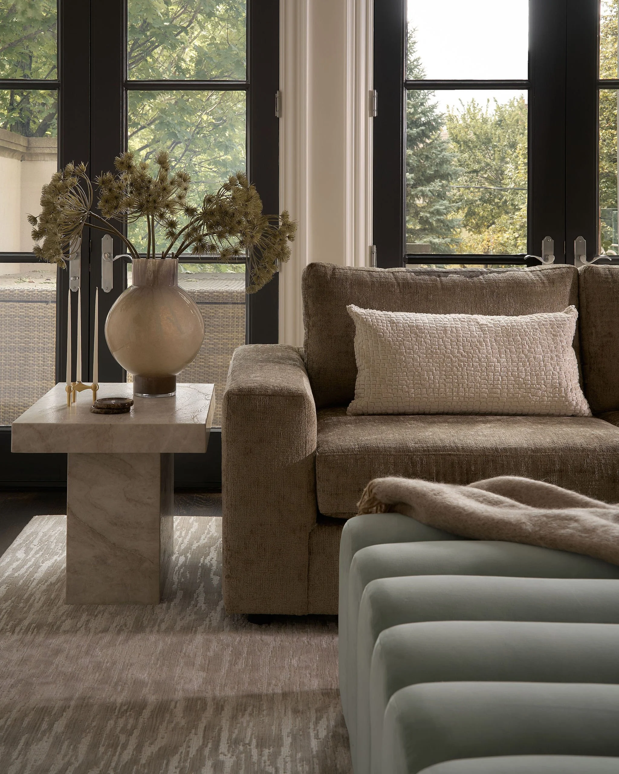 Living room with beige sofa, white textured pillow, marble coffee table with a vase of dried plants, and large windows showing green trees outside.