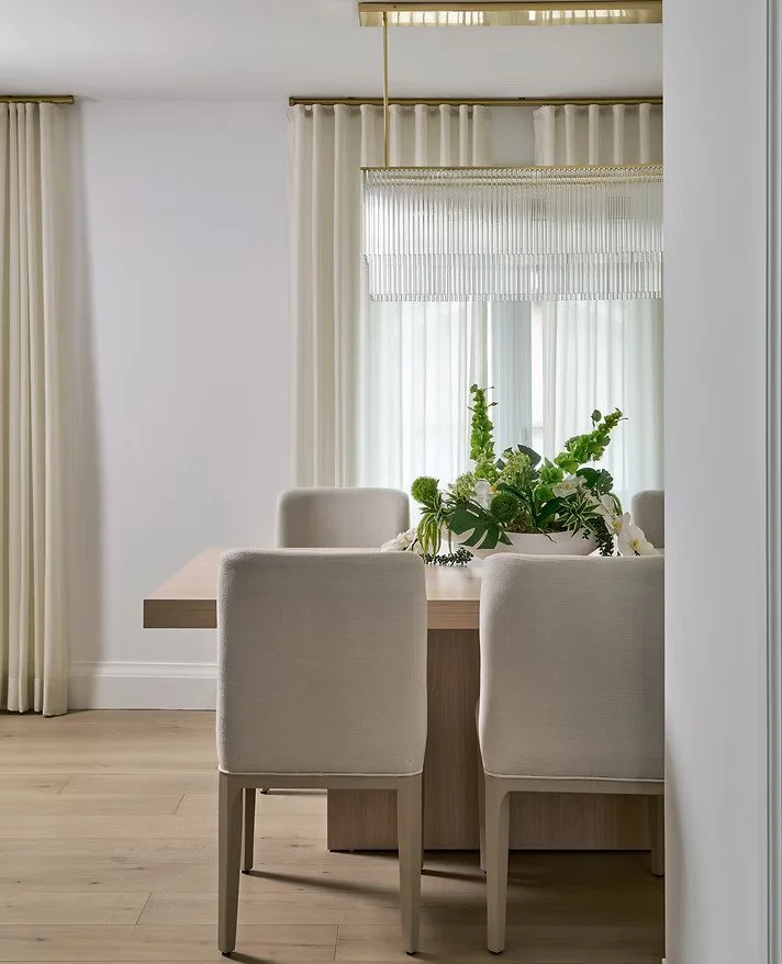 Dining room with a wooden table, four beige upholstered chairs, a potted plant centerpiece, and cream curtains on windows