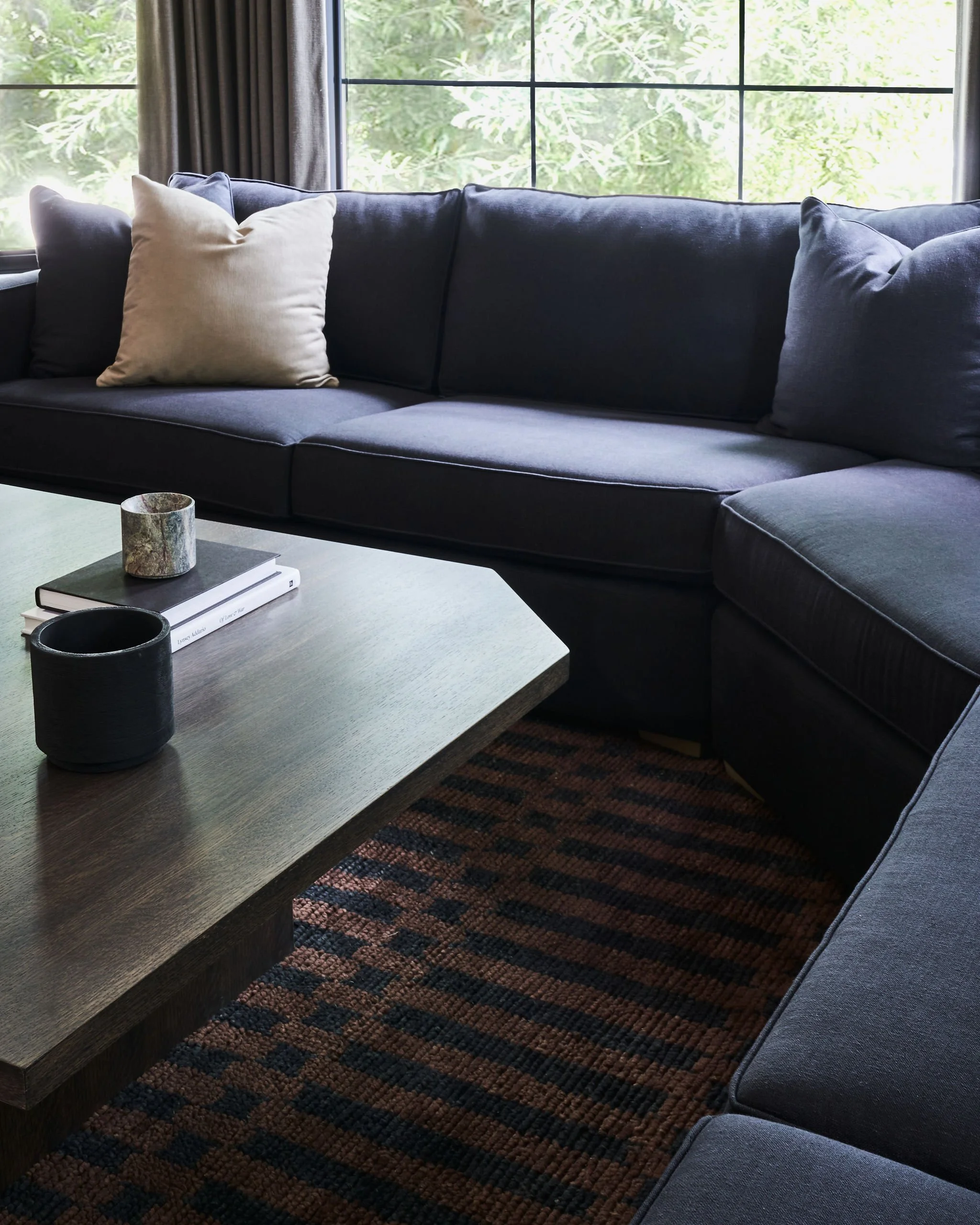 A black sofa with a beige pillow in a room with large windows showing green foliage outside. A wooden coffee table in front of the sofa has a black cup, two books, and a candle holder.