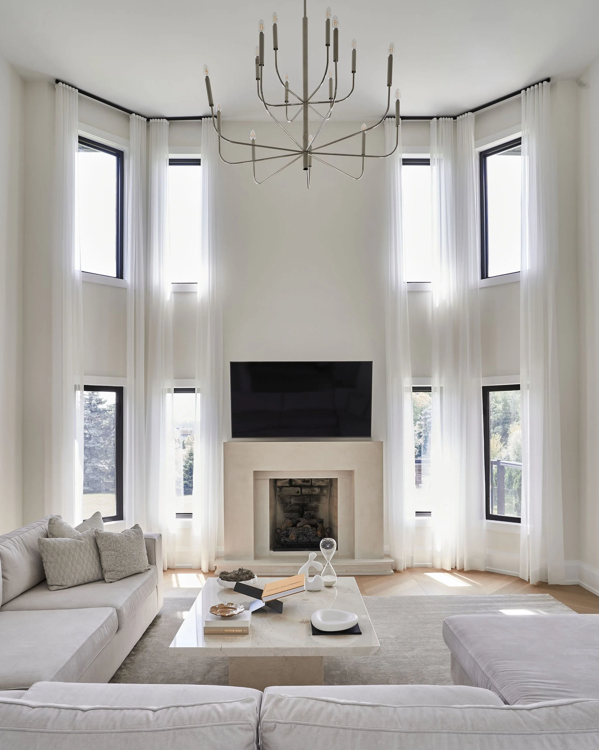 Bright living room with tall windows, white curtains, a modern chandelier, a fireplace, a large flat-screen TV, a light-colored sofa with cushions, a marble coffee table with decorative objects, and a neutral-colored area rug.
