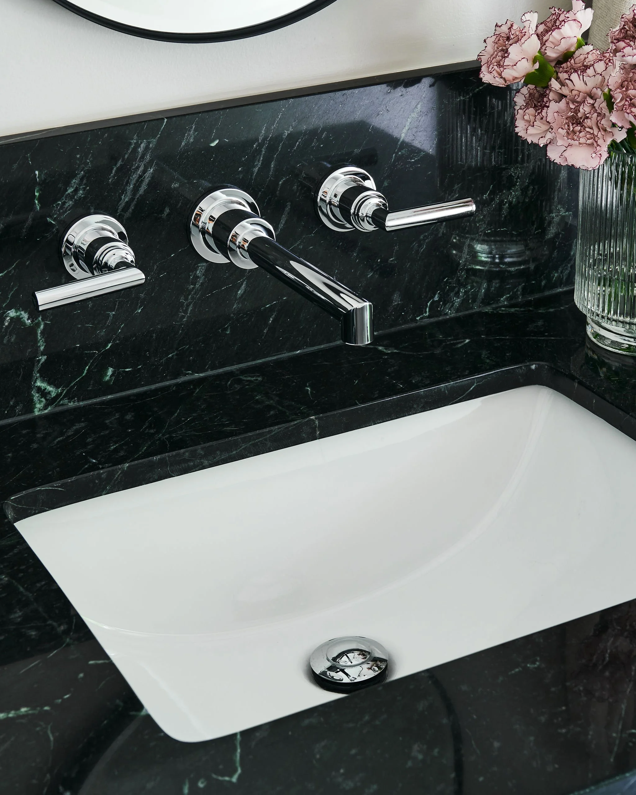 Close-up of a bathroom sink with a green marble countertop, a white ceramic basin, and a chrome faucet. A glass vase with pink and purple carnations is on the right side of the sink.