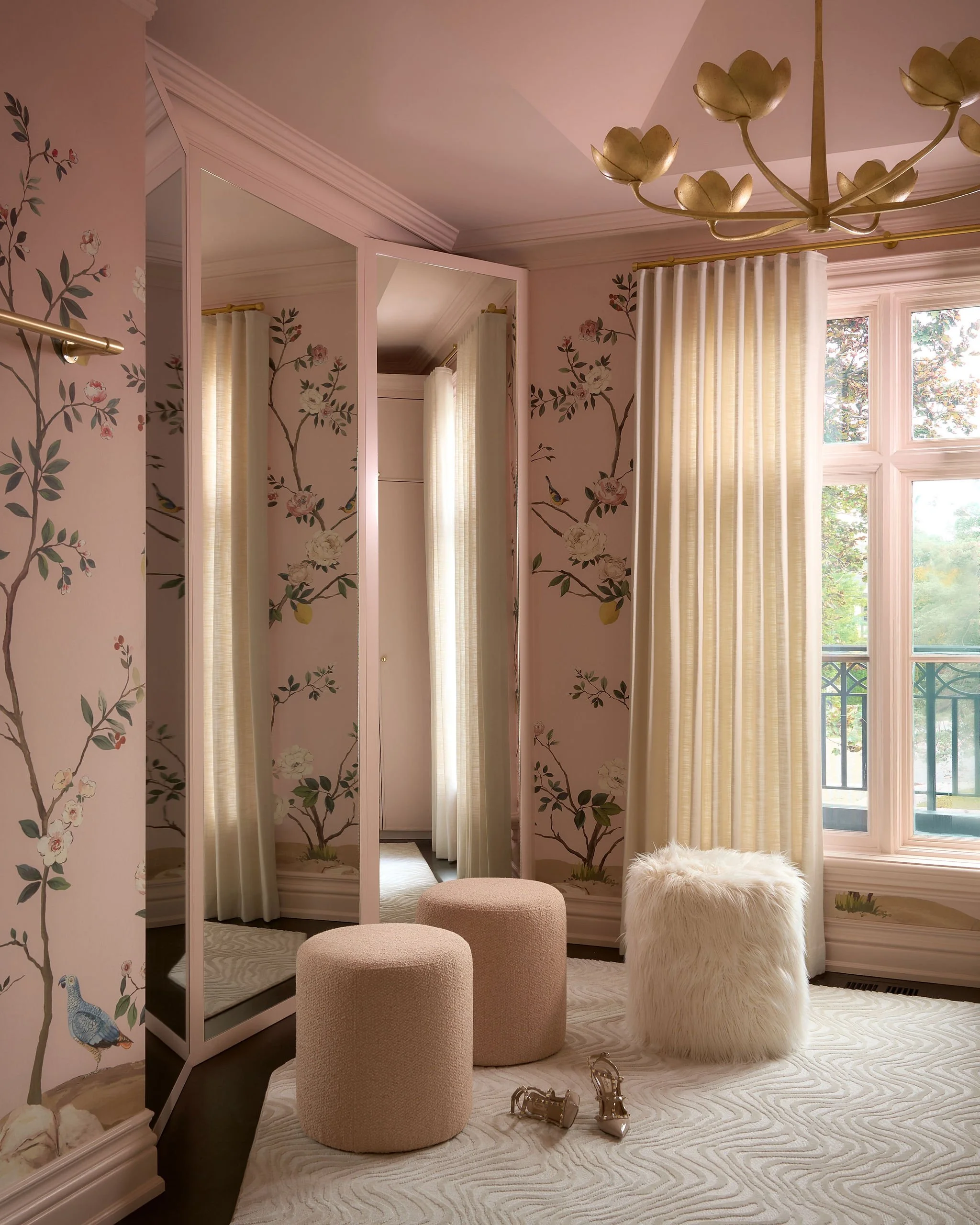 A cozy dressing area with floral wallpaper, a mirror wardrobe, cream-colored curtains, a window showing trees outside, and plush seating including a furry ottoman and two upholstered stools. A pair of high-heeled shoes is on the rug.