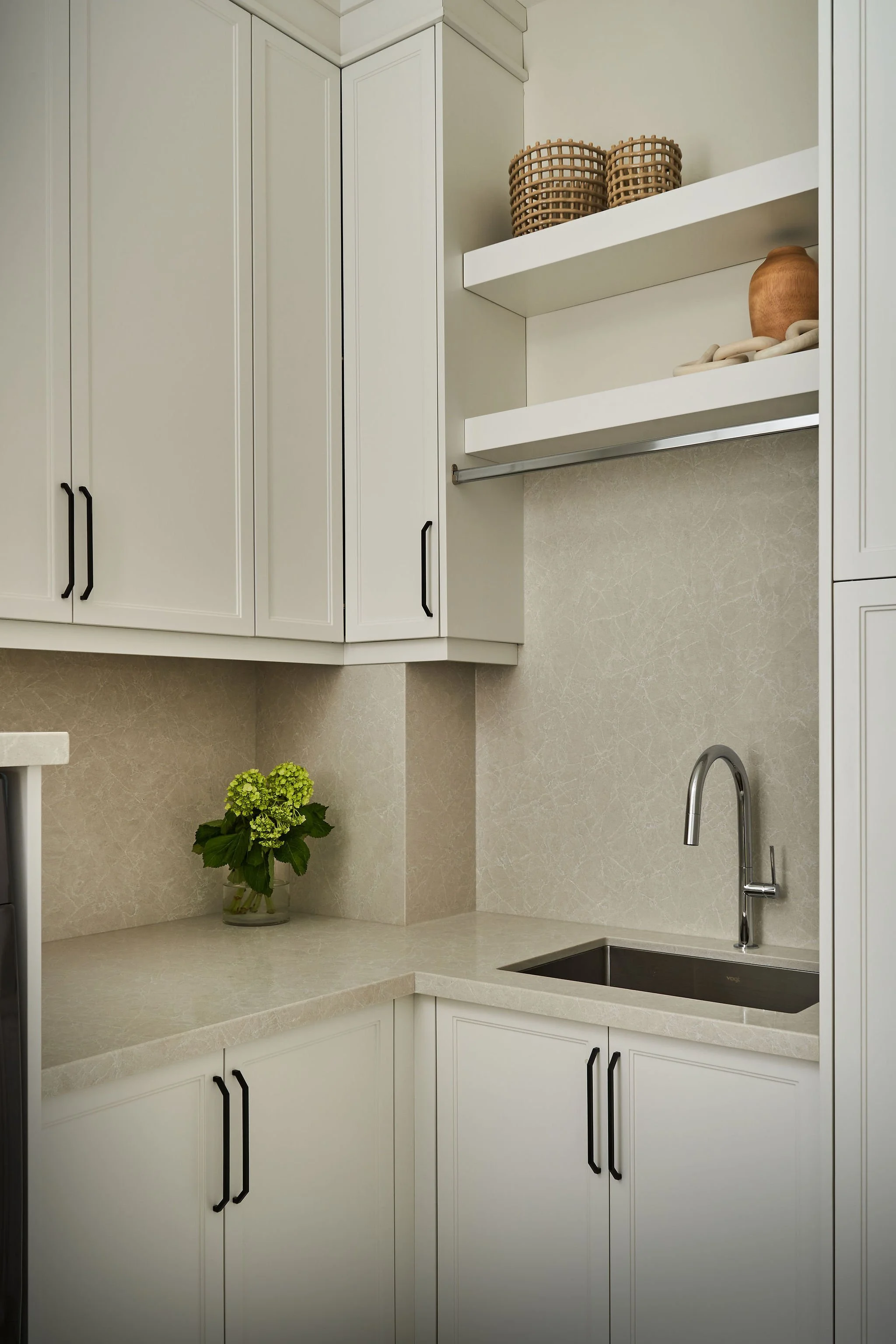 Clean white kitchen with black cabinet handles, a beige countertop, a stainless steel faucet over a black sink, a potted green plant, and open shelves with woven storage baskets and ceramic vases.