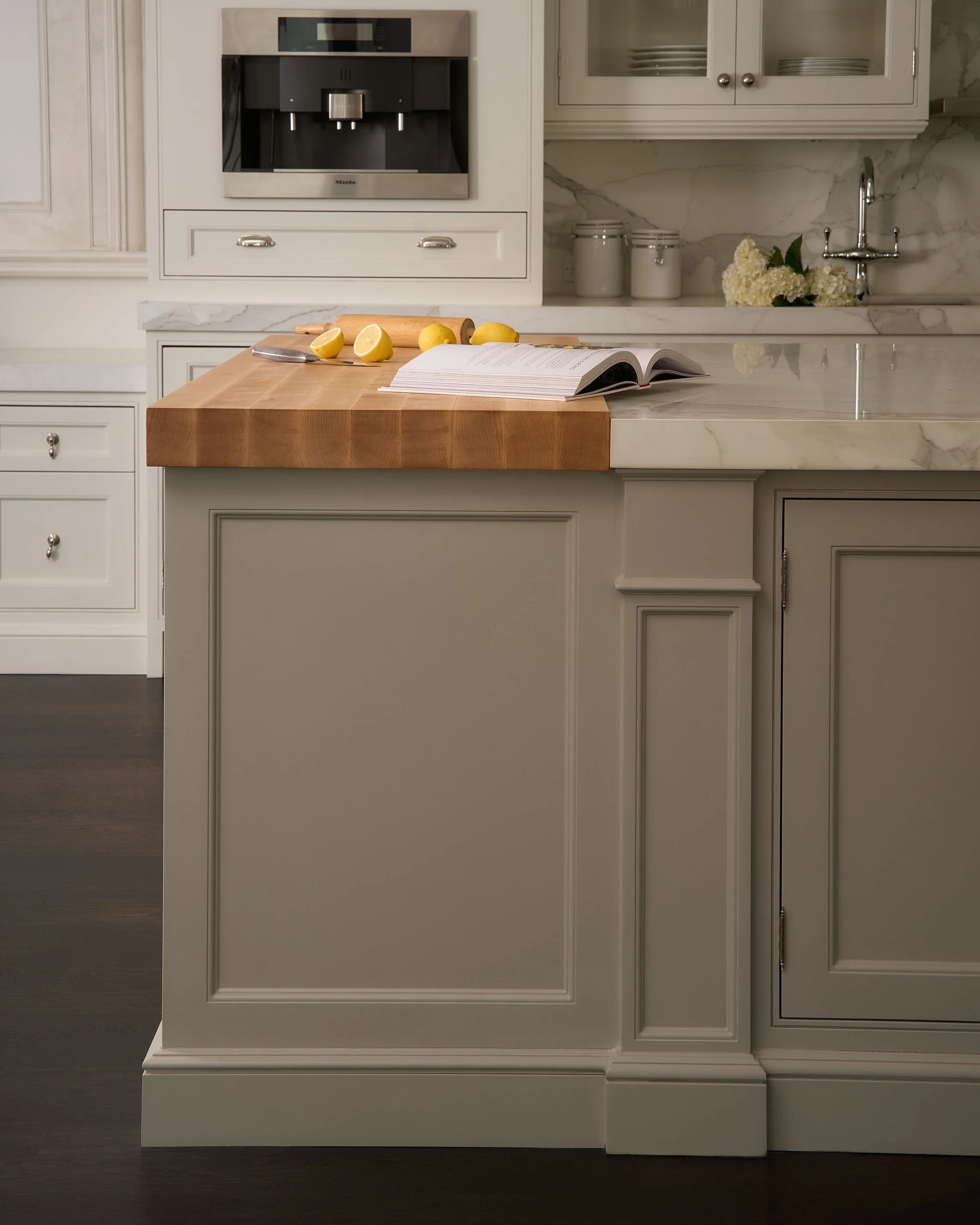 Kitchen island with a wooden cutting board, lemon halves, an open cook book, white cabinets, and a marble countertop. In the background, a coffee machine, plates, jars, and a flower arrangement are visible.