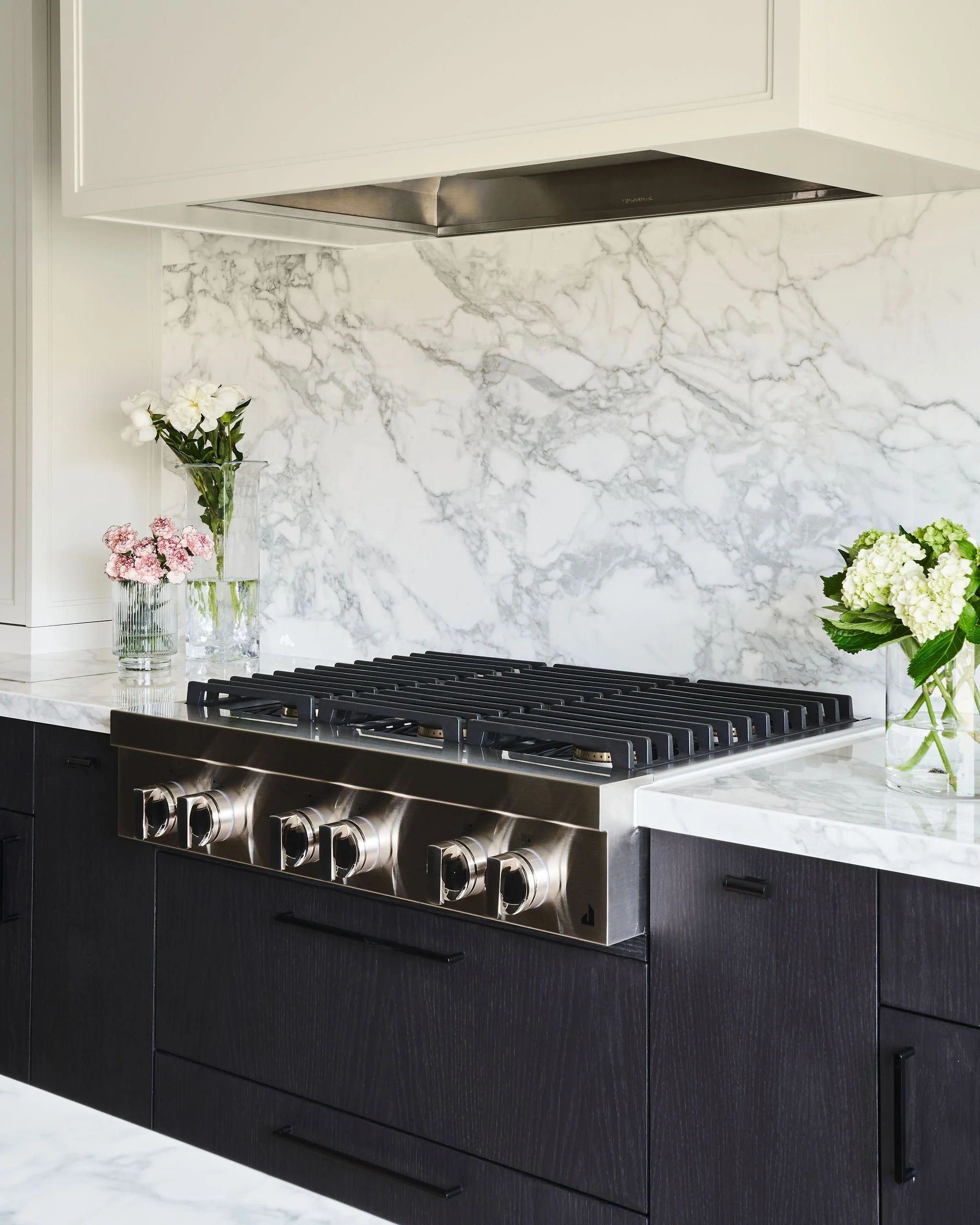 Modern kitchen with white marble countertops, black cabinets, stainless steel stovetop, and floral arrangements in glass vases.