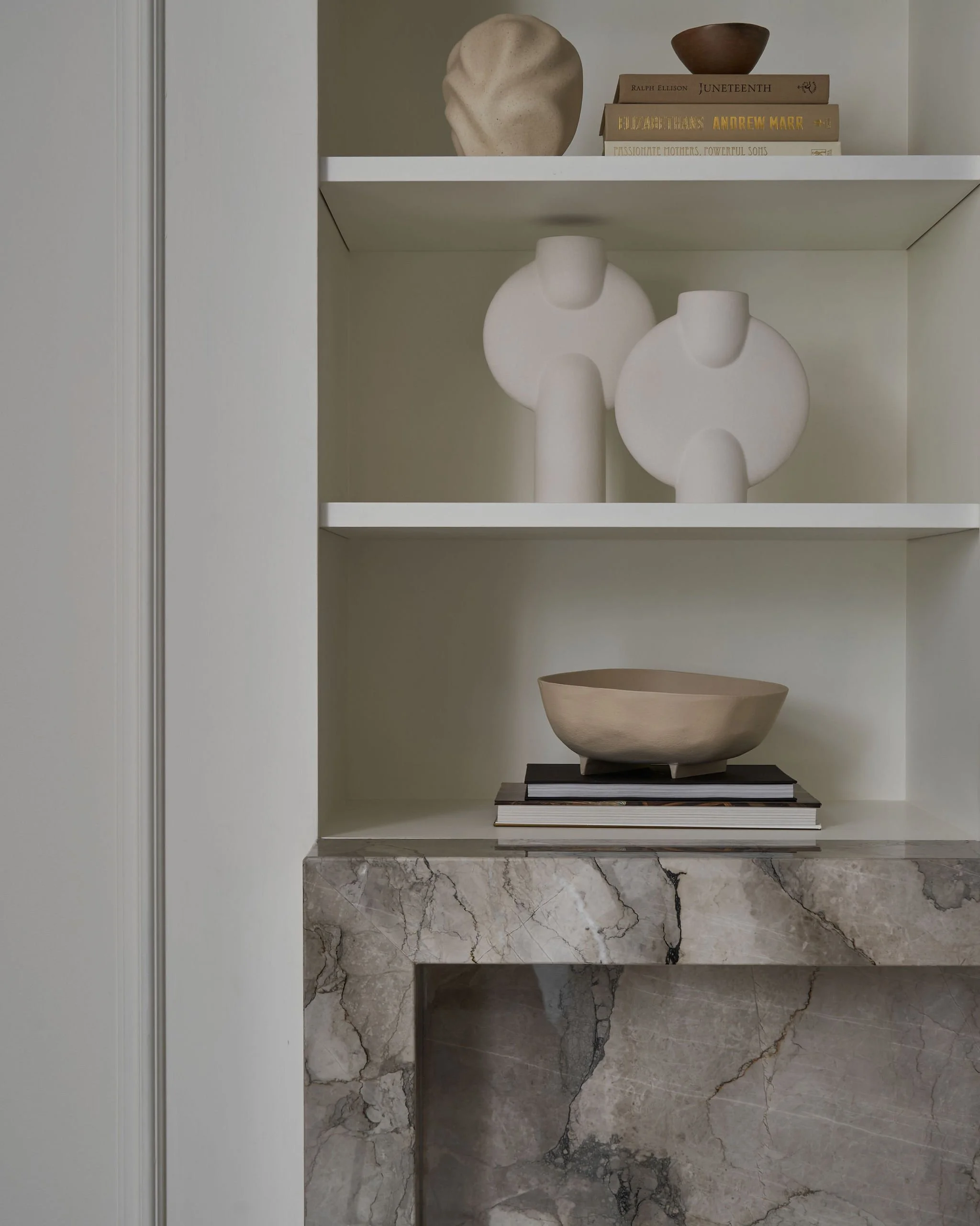 Open shelf displaying decorative and functional items including a beige vase, a small brown bowl, several books, white ceramic sculptures, and a beige bowl on a black stand, all against a white wall with marble flooring.