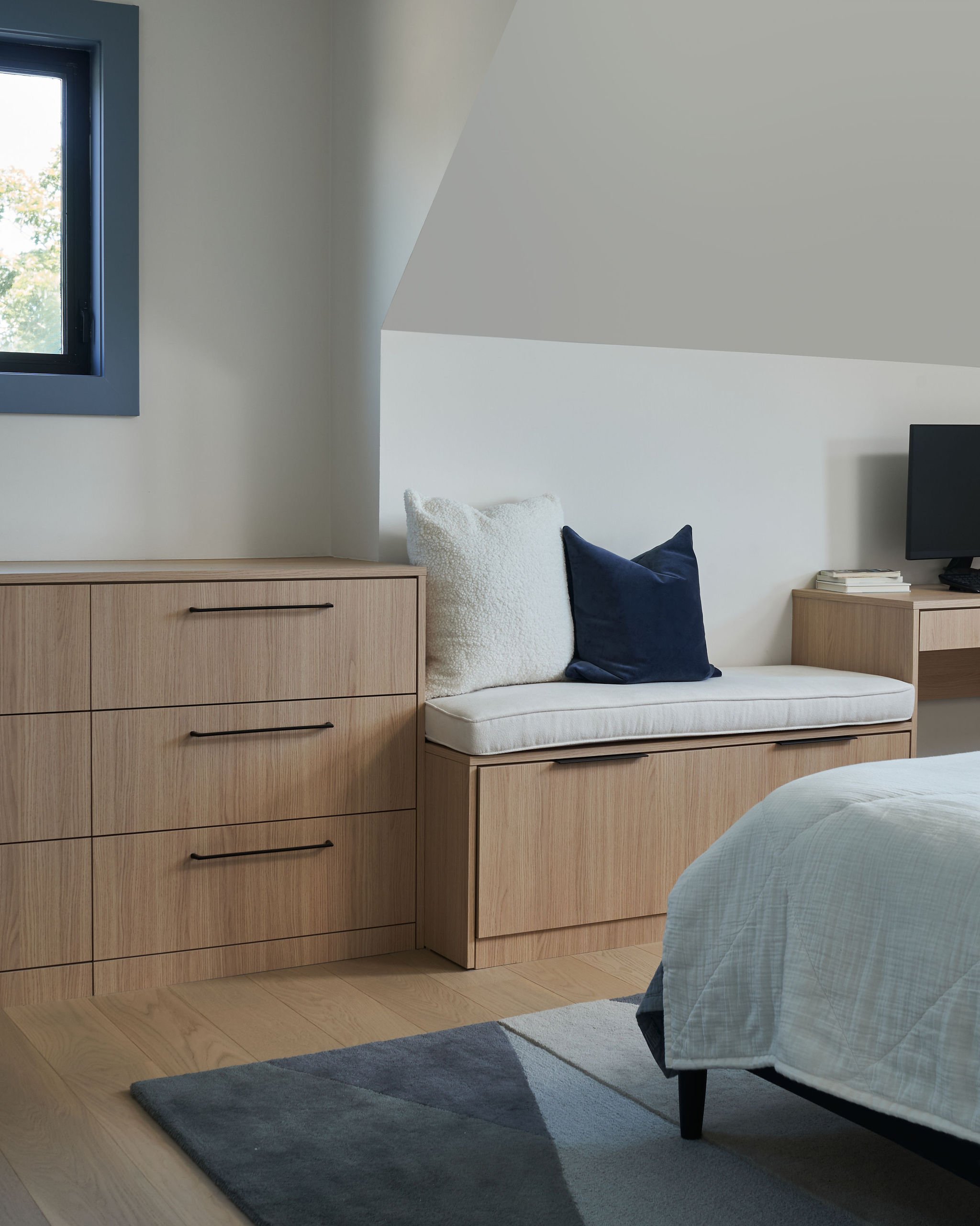 A modern bedroom with a wooden dresser, a white sofa with pillows, a bed with a light blue quilt, a gray and blue rug, a window with a black frame, a small side table with books and a TV.