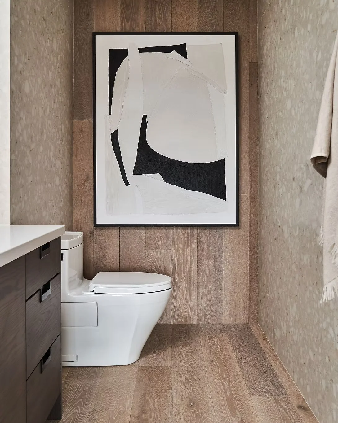 Modern bathroom with a white toilet, dark wooden cabinet, and a large abstract black and white painting on a wood-paneled wall.